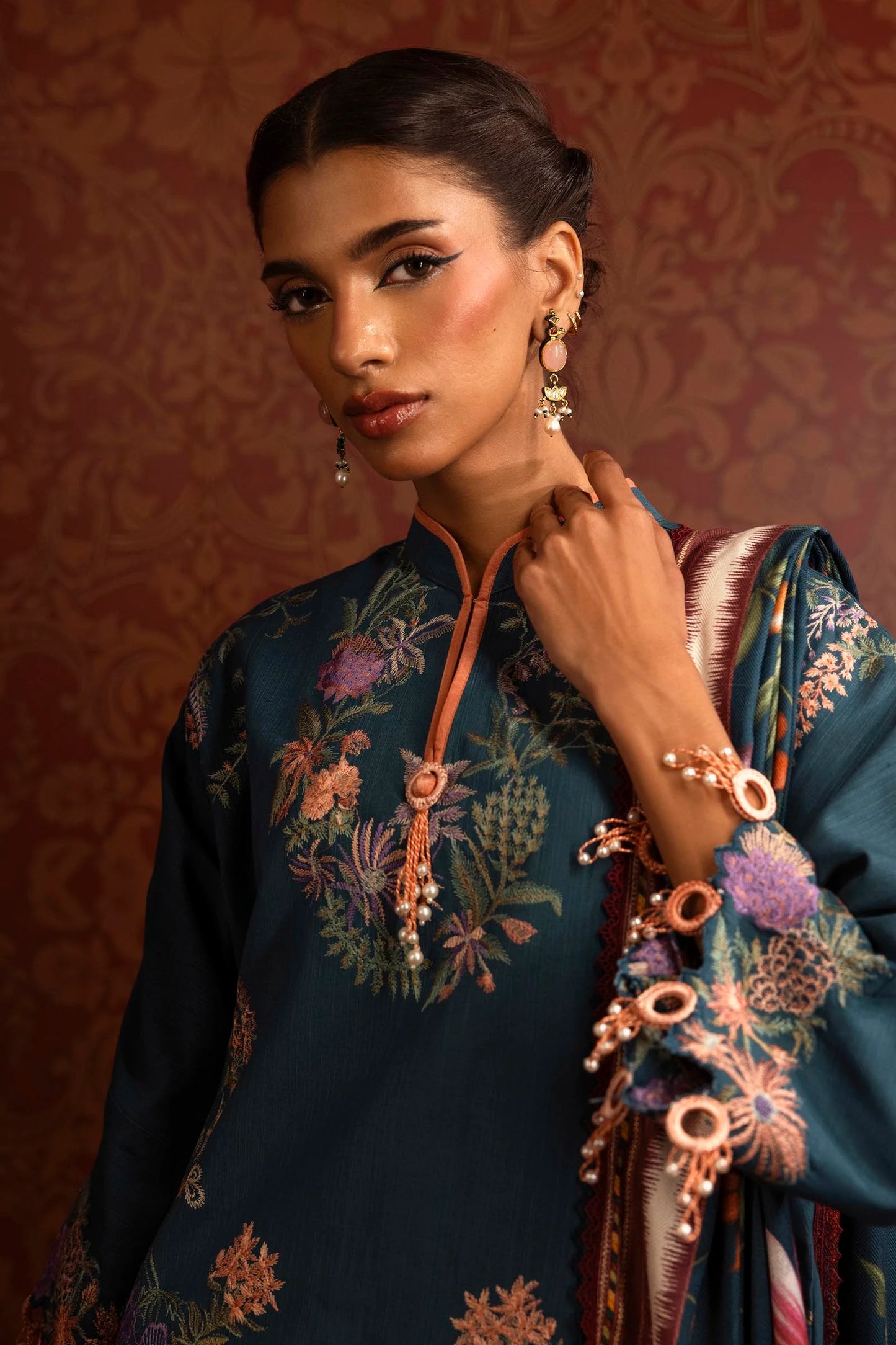 A model wears a three-piece outfit from the Sana Safinaz Muzlin Winter '24 Collection. The ensemble features an embroidered front kali on slub fabric, complemented by a dyed slub back and sleeves with intricate embroidery. A vibrant digital printed shawl adds elegance, while dyed cotton pants complete the look. Perfect for women seeking premium Pakistani designer clothing. Available online in the UK.