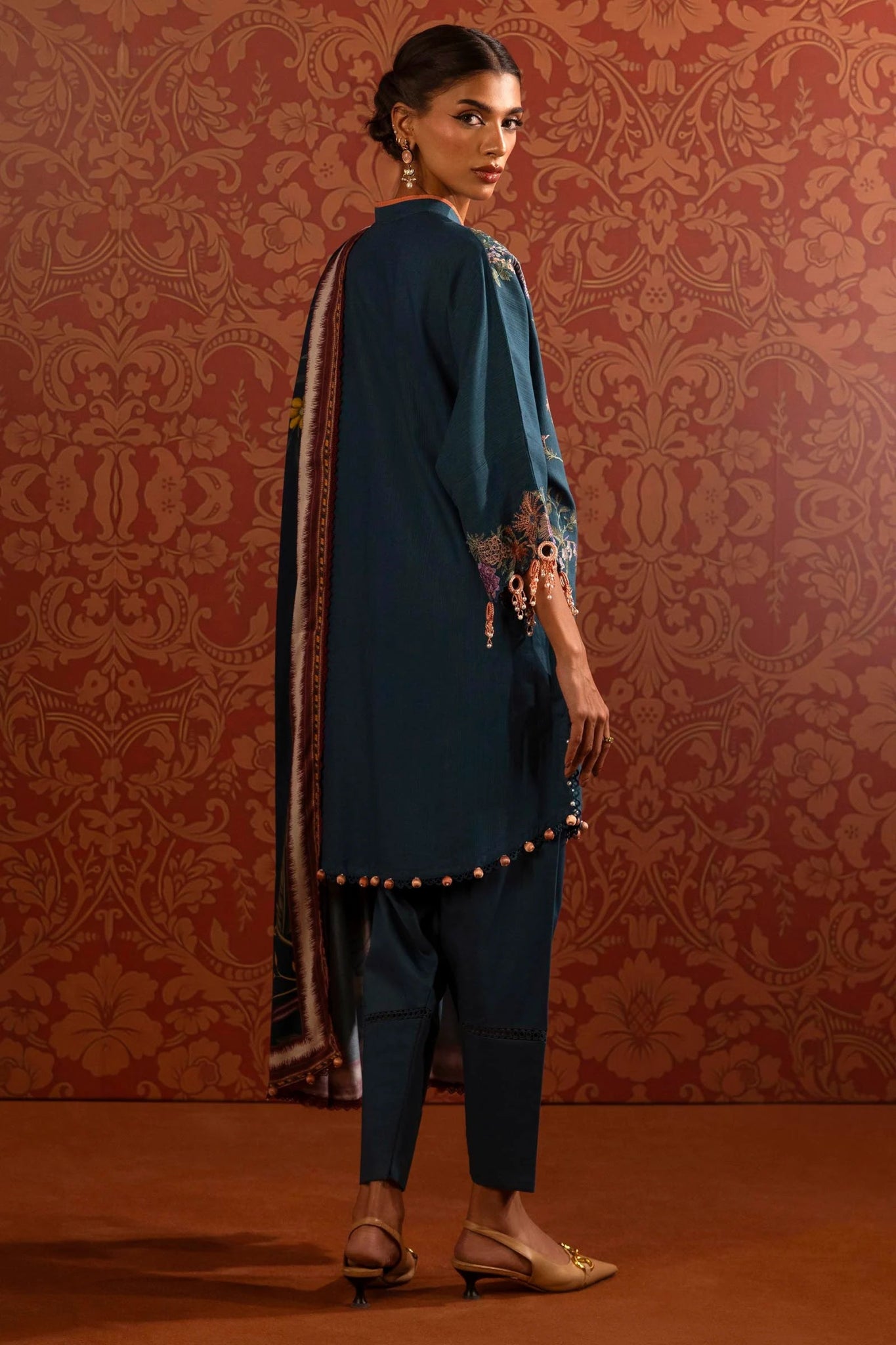 A model wears a three - piece outfit from the Sana Safinaz Muzlin Winter '24 Collection. The ensemble features an embroidered front kali on slub fabric, complemented by a dyed slub back and sleeves with intricate embroidery. A vibrant digital printed shawl adds elegance, while dyed cotton pants complete the look. Perfect for women seeking premium Pakistani designer clothing. Available online in the UK.