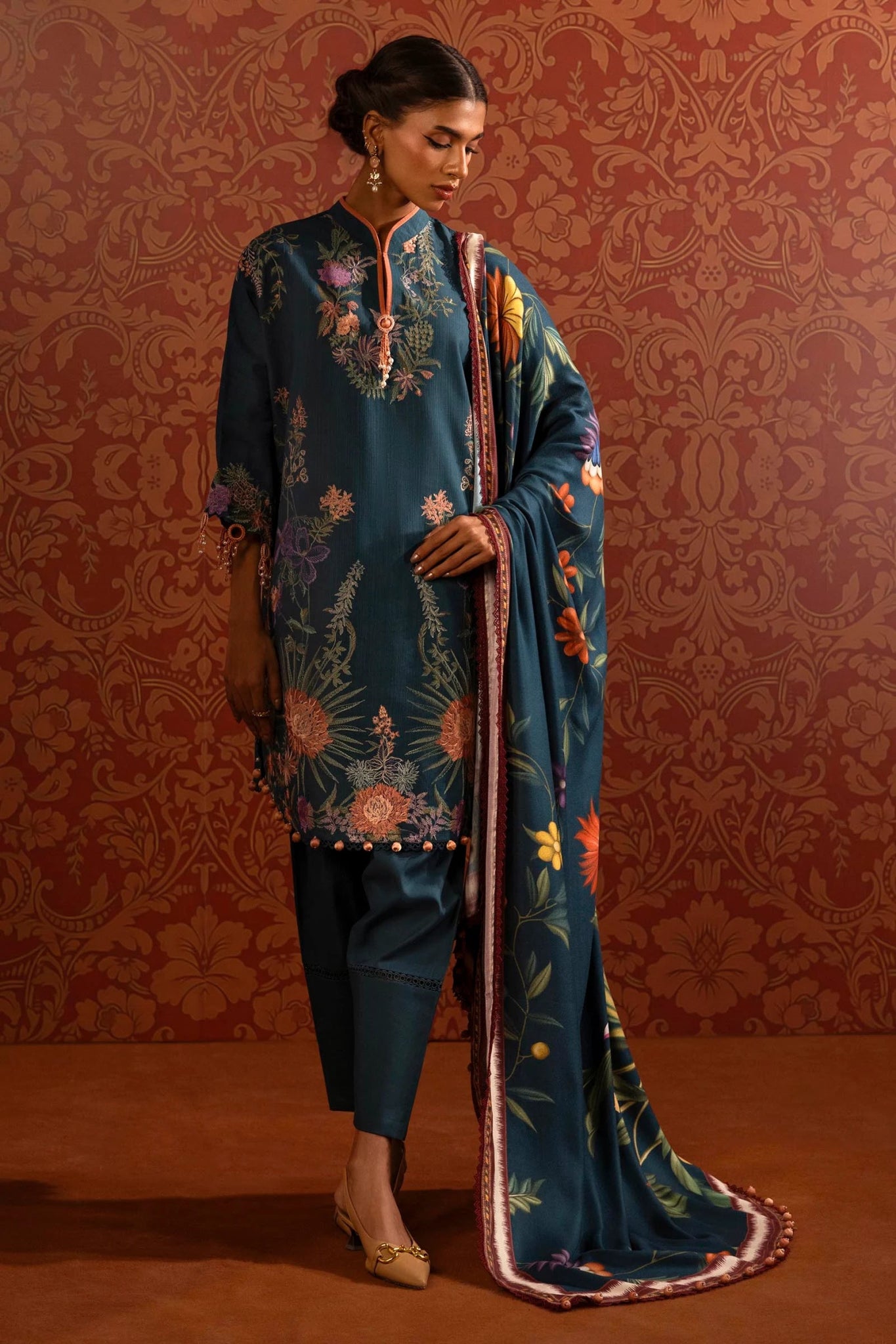 A model wears a three - piece outfit from the Sana Safinaz Muzlin Winter '24 Collection. The ensemble features an embroidered front kali on slub fabric, complemented by a dyed slub back and sleeves with intricate embroidery. A vibrant digital printed shawl adds elegance, while dyed cotton pants complete the look. Perfect for women seeking premium Pakistani designer clothing. Available online in the UK.