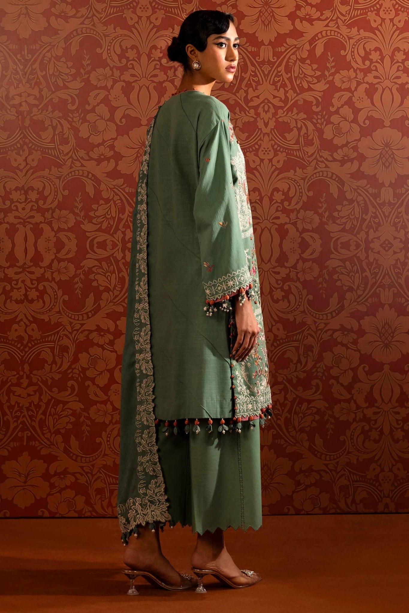 A model wears a three - piece outfit from the Sana Safinaz Muzlin Winter '24 Collection. It includes an embroidered front kali on slub fabric, complemented by a dyed slub back and intricately embroidered sleeves. The look is finished with an elegant embroidered shawl featuring detailed pallus, and dyed cotton pants, creating a refined and cohesive ensemble. Ideal for those seeking luxurious Pakistani fashion. Available online.