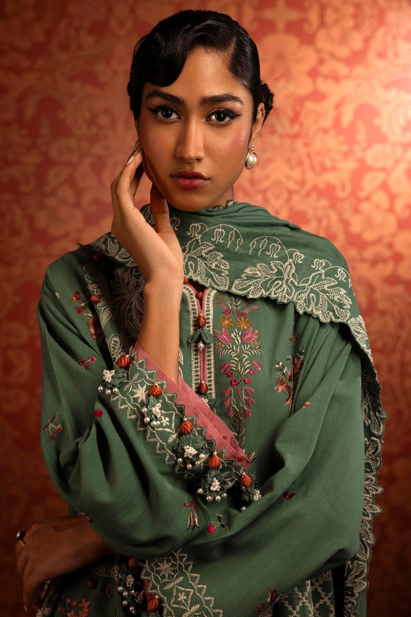 A model wears a three - piece outfit from the Sana Safinaz Muzlin Winter '24 Collection. It includes an embroidered front kali on slub fabric, complemented by a dyed slub back and intricately embroidered sleeves. The look is finished with an elegant embroidered shawl featuring detailed pallus, and dyed cotton pants, creating a refined and cohesive ensemble. Ideal for those seeking luxurious Pakistani fashion. Available online.