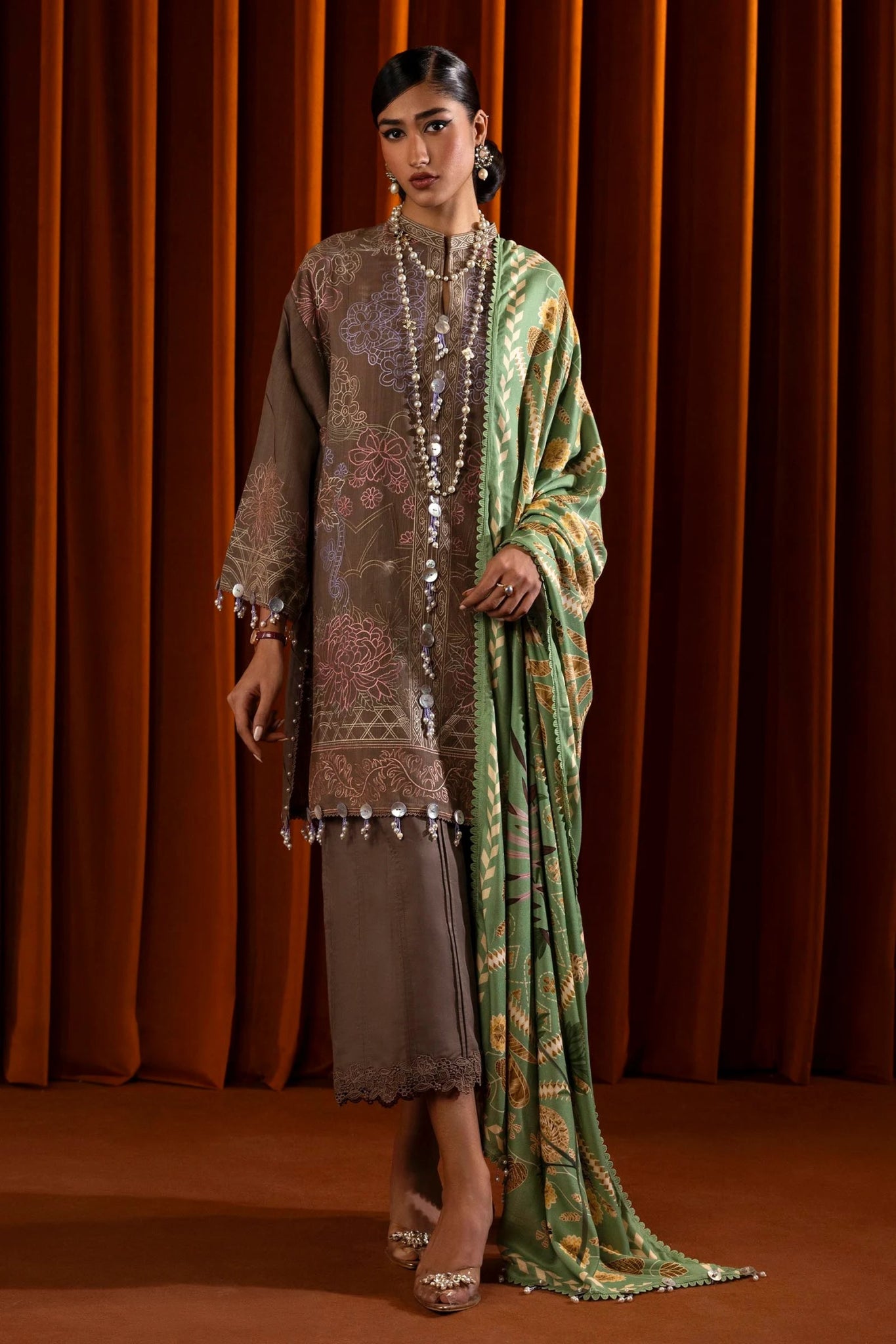 A model is wearing a three - piece outfit from the Sana Safinaz Muzlin Winter '24 Collection. The ensemble features an embroidered front kali on slub fabric, paired with a dyed slub back and sleeves, all detailed with delicate embroidery. It includes a digitally printed shawl with intricate patterns, complemented by dyed cotton pants. The luxurious and sophisticated design, with its attention to detail, offers a chic choice for women seeking elegant Pakistani designer clothing. Available online