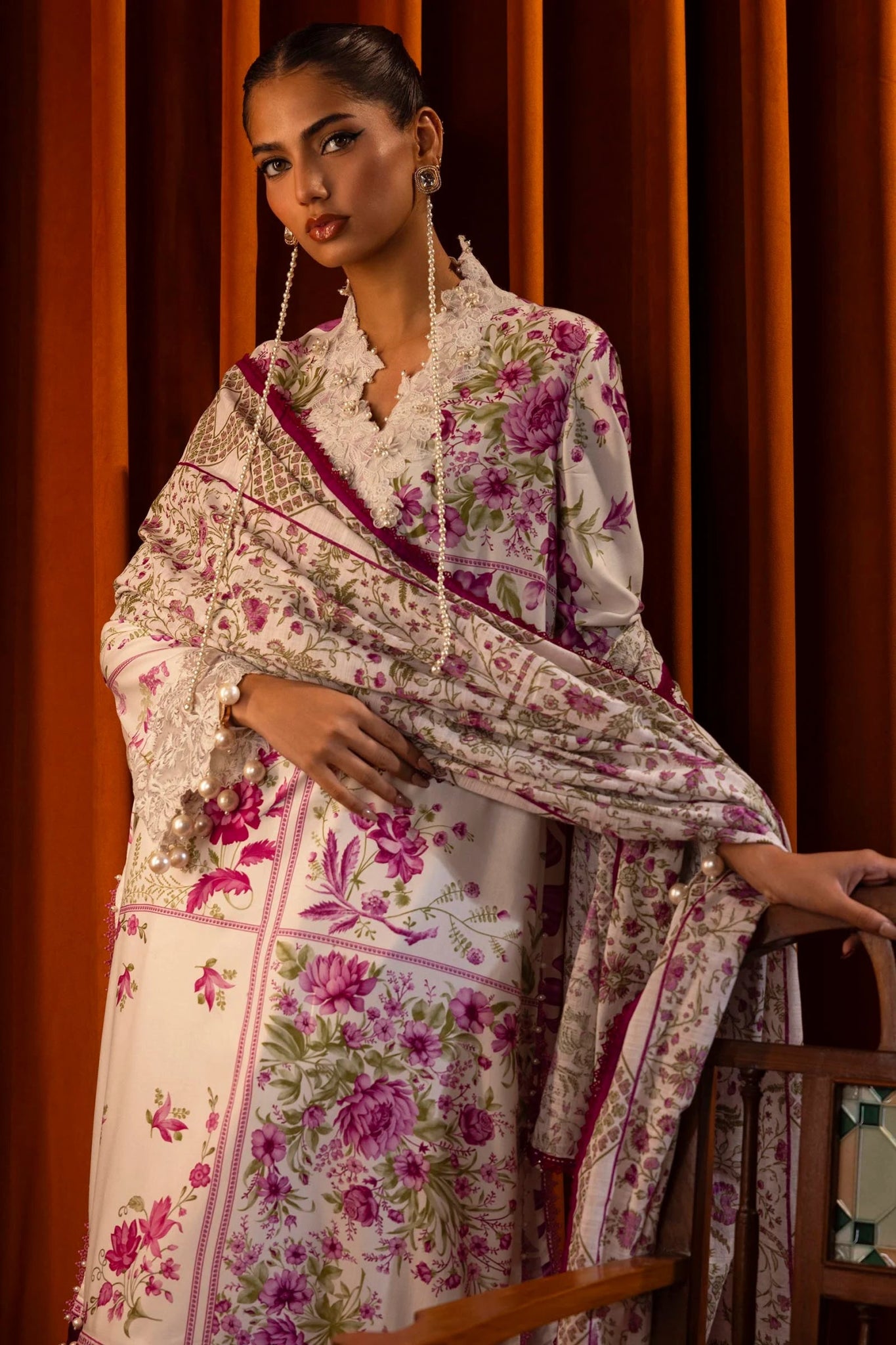 Model wearing a 3 - piece floral suit in white and pink from the Sana Safinaz Muzlin Winter 2024 collection. The outfit features a digitally printed linen front, back, and sleeves, with embroidered organza patti for the neckline and sleeves. Styled with a printed khaddar dupatta and dyed cotton pants.