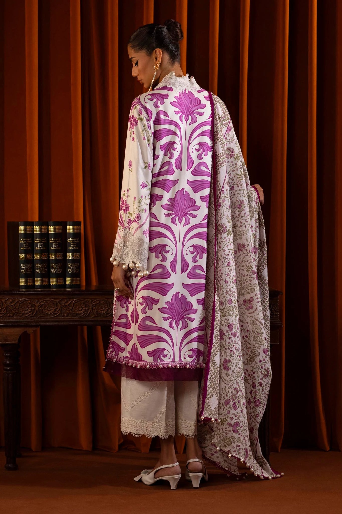 Model wearing a 3 - piece floral suit in white and pink from the Sana Safinaz Muzlin Winter 2024 collection. The outfit features a digitally printed linen front, back, and sleeves, with embroidered organza patti for the neckline and sleeves. Styled with a printed khaddar dupatta and dyed cotton pants.