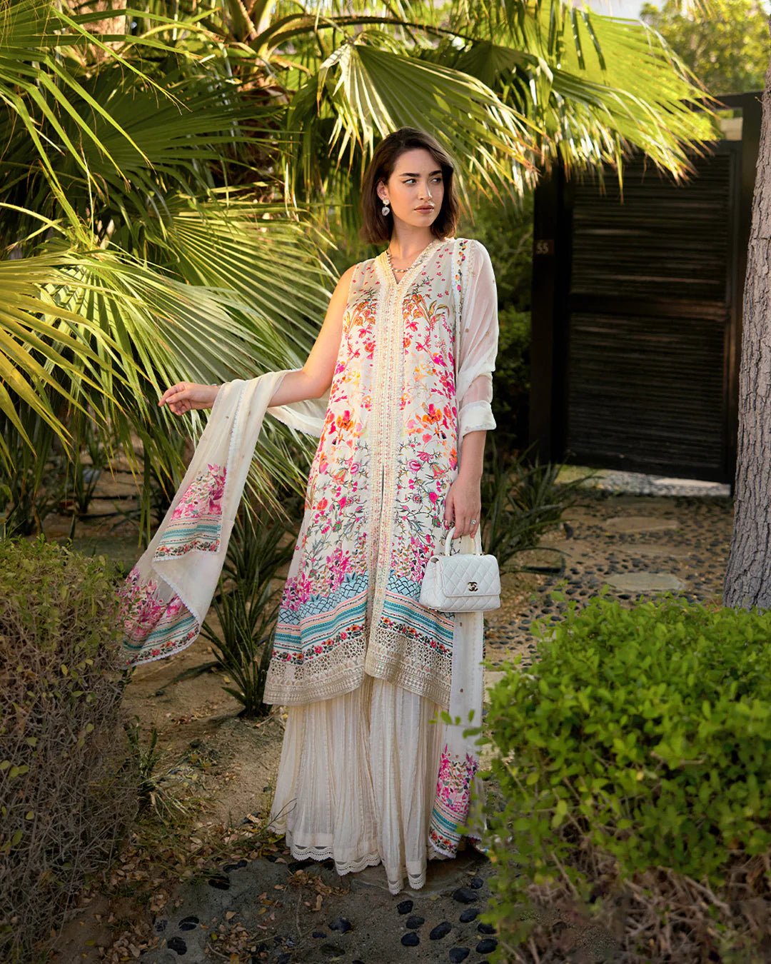 Model wearing Faiza Saqlain’s Beldene from the Carmela collection, featuring a cream embroidered silk shirt with floral patterns, paired with a crushed silk sharara and printed chiffon dupatta. Perfect for Pakistani clothes UK.