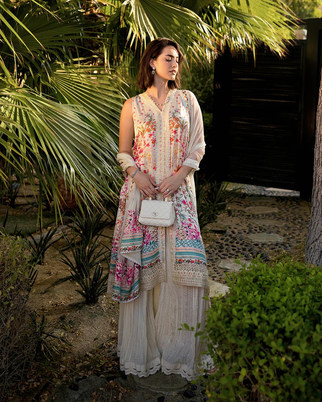 Model wearing Faiza Saqlain’s Beldene from the Carmela collection, featuring a cream embroidered silk shirt with floral patterns, paired with a crushed silk sharara and printed chiffon dupatta. Perfect for Pakistani clothes UK.