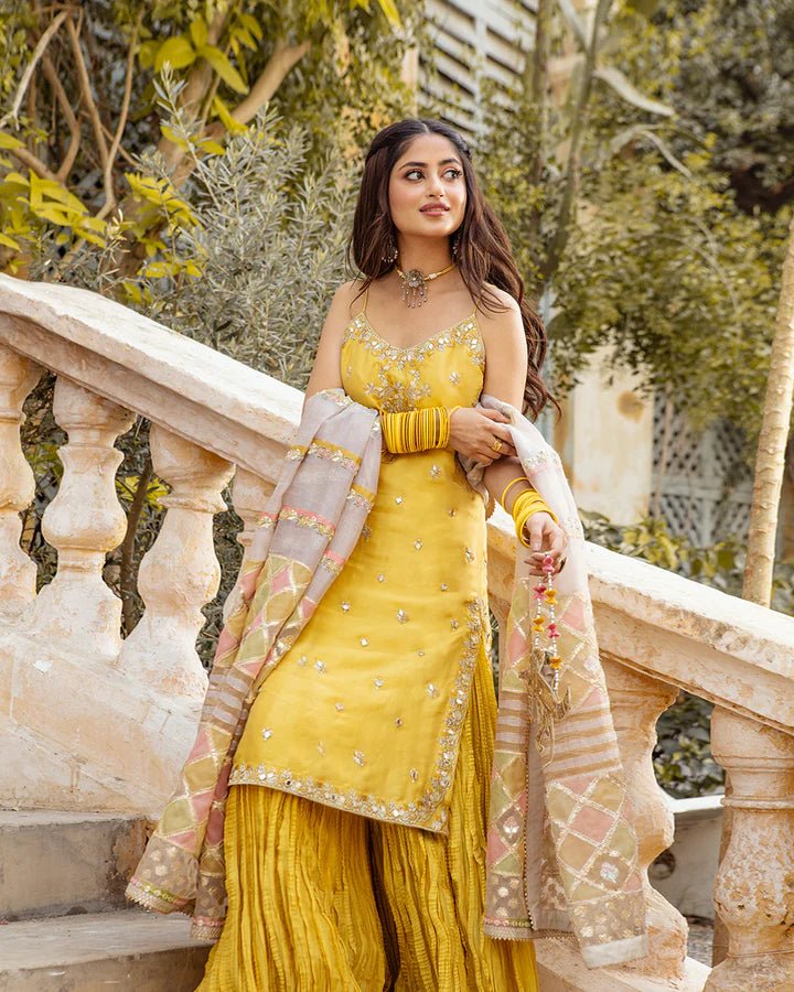 Model wearing Faiza Saqlain Fara dress in yellow mehndi dress from the Dhoop Kinaray Formals collection, ideal for weddings, available for Pakistani clothes online in the UK.