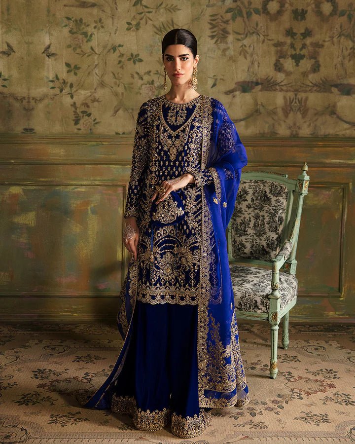 Model wearing a royal blue Seyhana dress with intricate gold embroidery from Faiza Saqlain's Luxe Formals Manaar Nov '23 collection. Pakistani clothes online in UK.