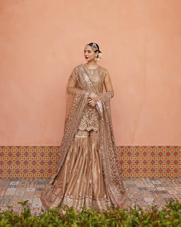 Model wearing a gold Jolina dress from Faiza Saqlain's Neorah Wedding Festive '24 Collection, featuring intricate embroidery and a flowing silhouette. Pakistani wedding clothes available online in the UK.