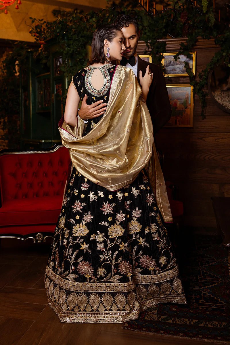 Model wearing Mushq DEJA VU - VELVET '24 Whispers of Yesterday, a black velvet gown with intricate gold and red floral embroidery, styled with a luxurious golden shawl. Ideal for winter weddings, available for Pakistani clothes online in the UK.