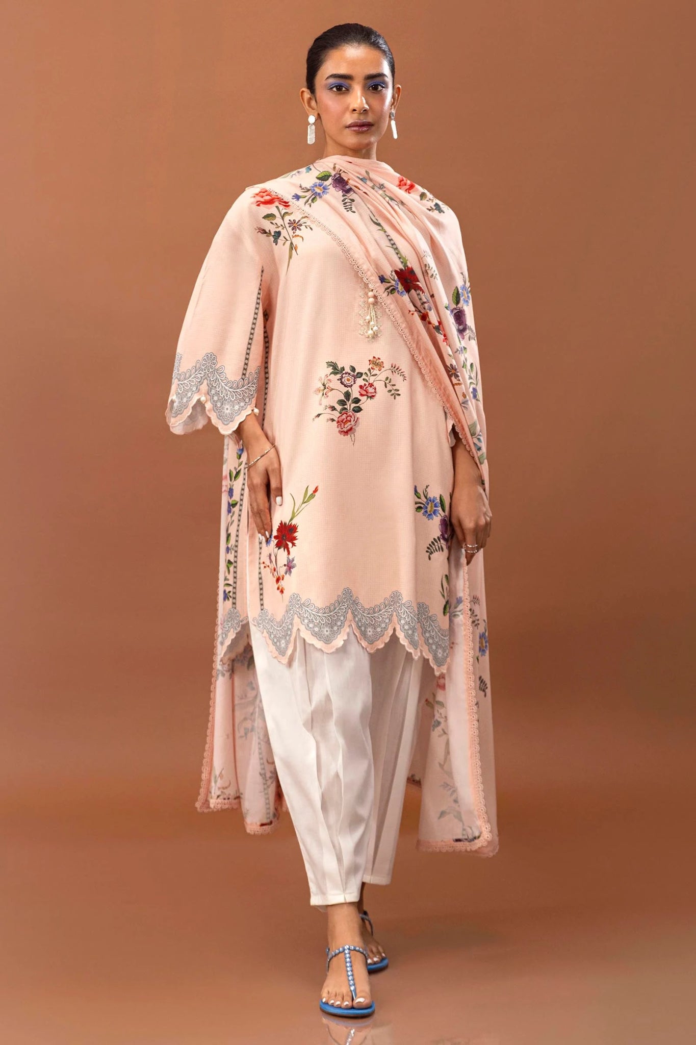 Model wearing a two - piece Sana Safinaz suit from Mahay Winter '24 in soft peach, featuring floral digital prints on a linen shirt, embroidered organza patti, and a matching printed dupatta. Available online in the UK.