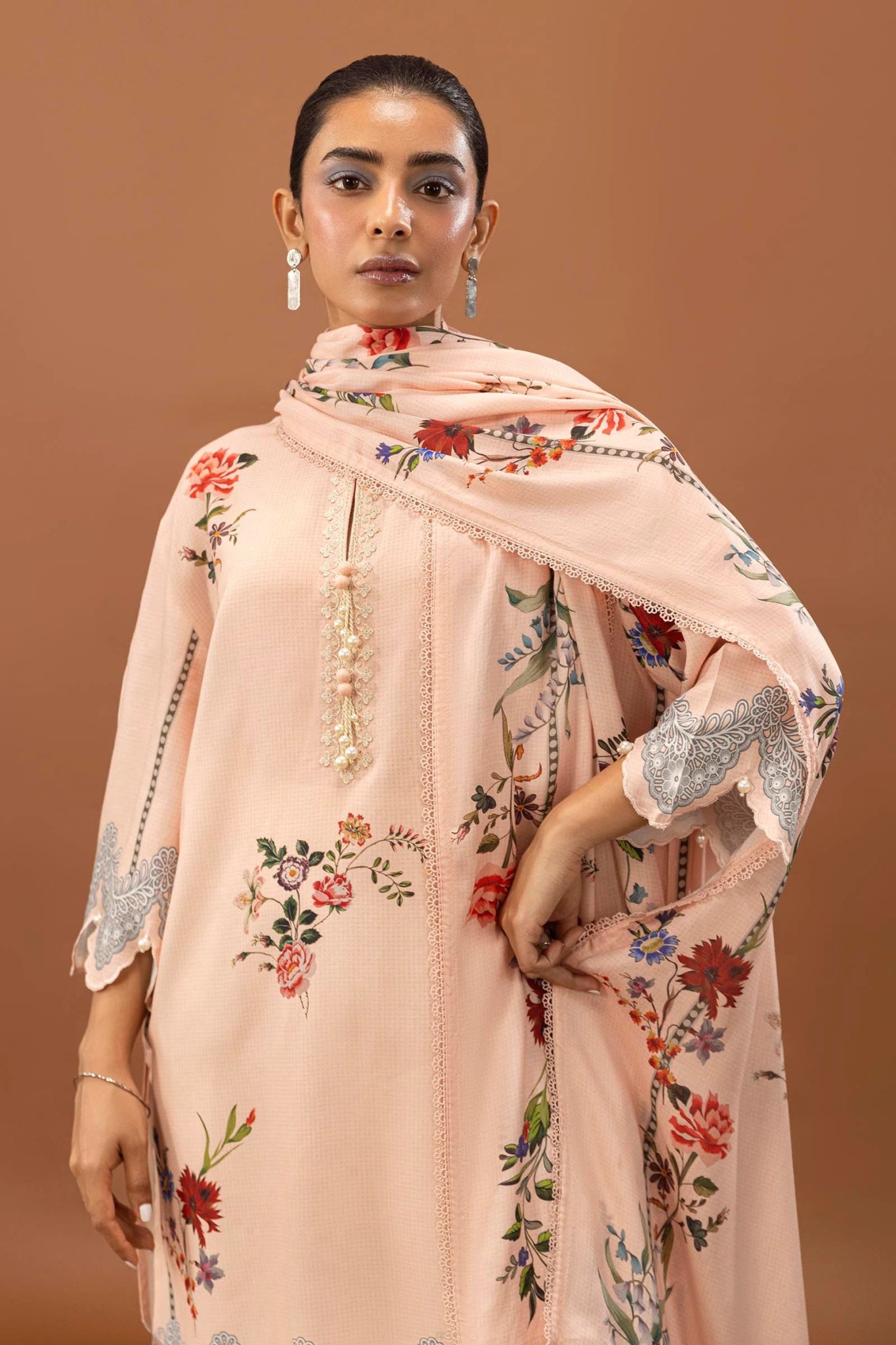 Model wearing a two - piece Sana Safinaz suit from Mahay Winter '24 in soft peach, featuring floral digital prints on a linen shirt, embroidered organza patti, and a matching printed dupatta. Available online in the UK.