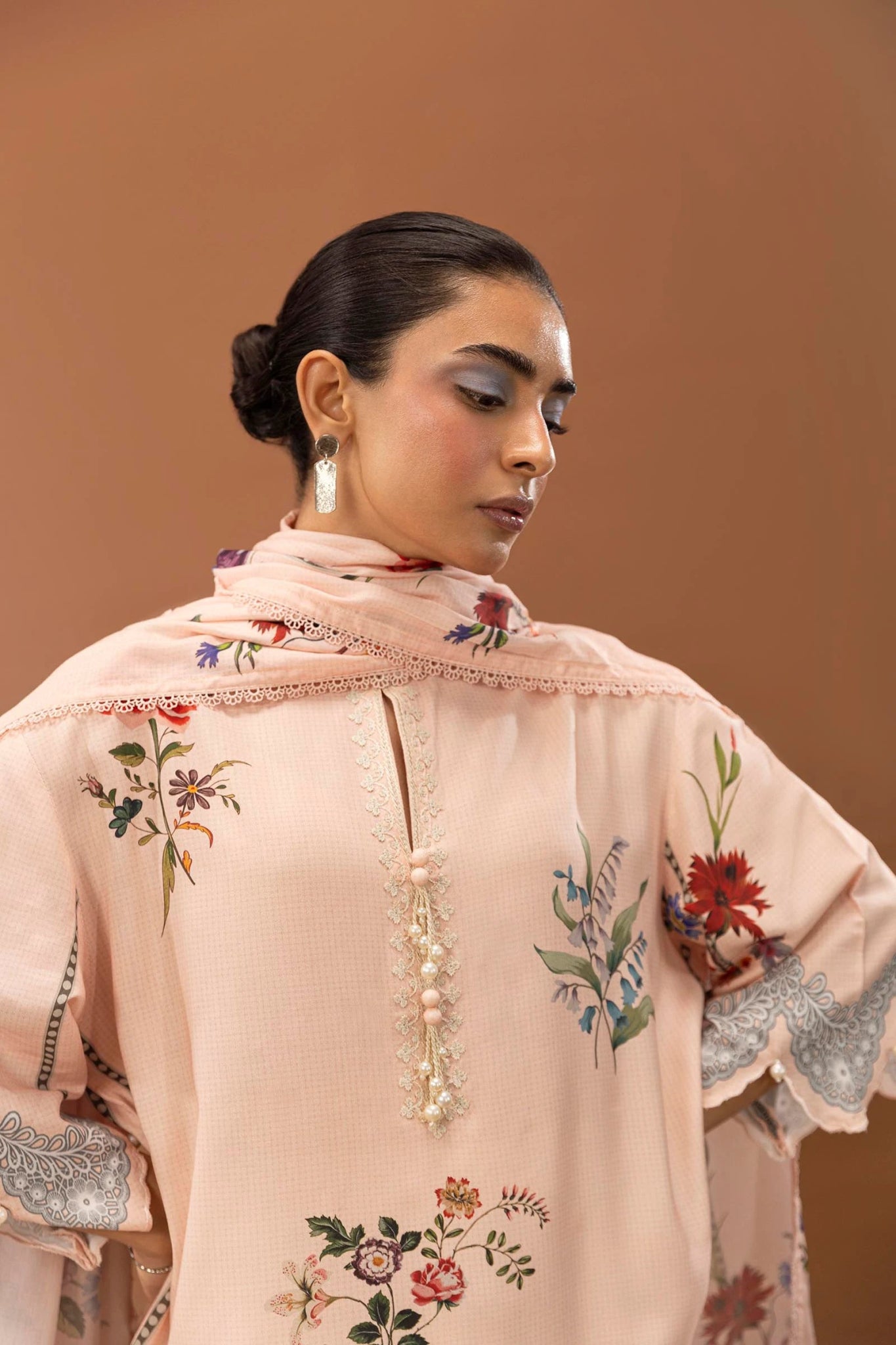 Model wearing a two - piece Sana Safinaz suit from Mahay Winter '24 in soft peach, featuring floral digital prints on a linen shirt, embroidered organza patti, and a matching printed dupatta. Available online in the UK.