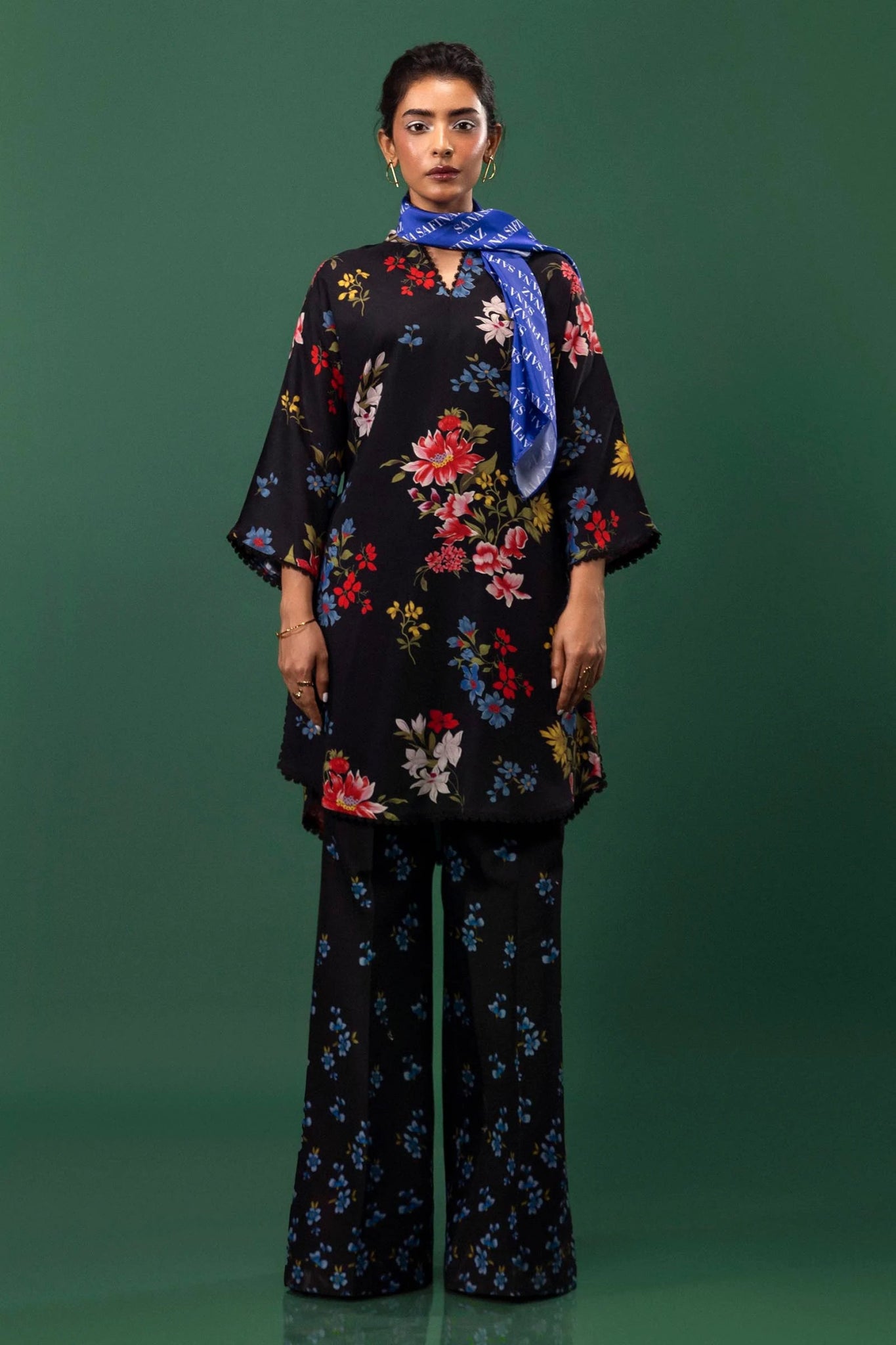 Model wearing a two - piece Sana Safinaz suit from Mahay Winter '24 featuring a black linen shirt with vibrant floral prints, embroidered organza accents, and matching printed cambric pants. Available online in the UK.