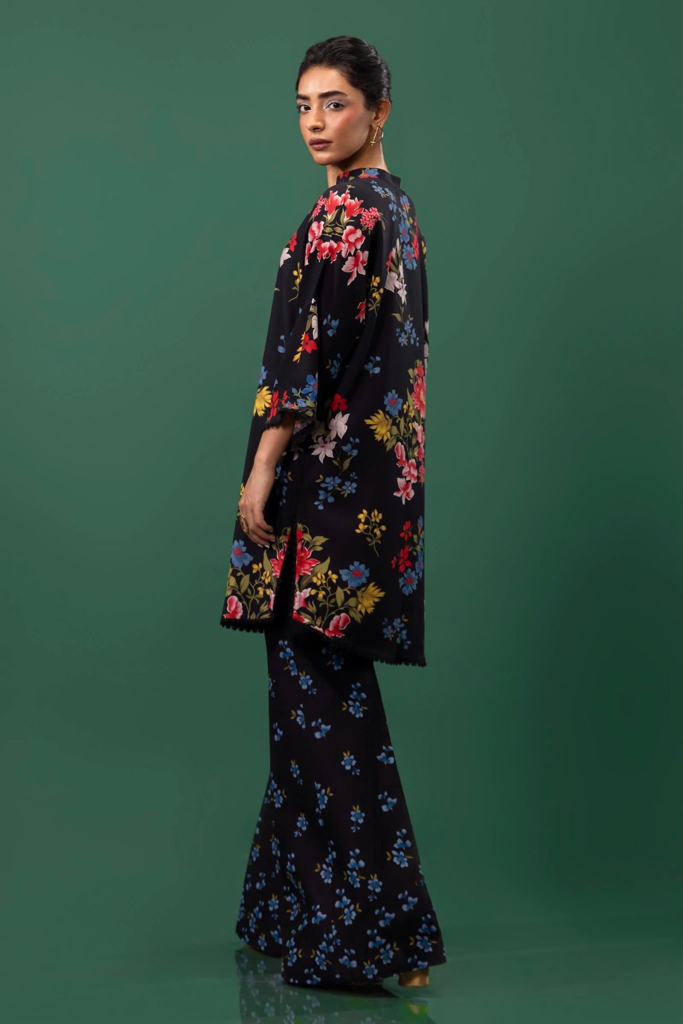 Model wearing a two - piece Sana Safinaz suit from Mahay Winter '24 featuring a black linen shirt with vibrant floral prints, embroidered organza accents, and matching printed cambric pants. Available online in the UK.