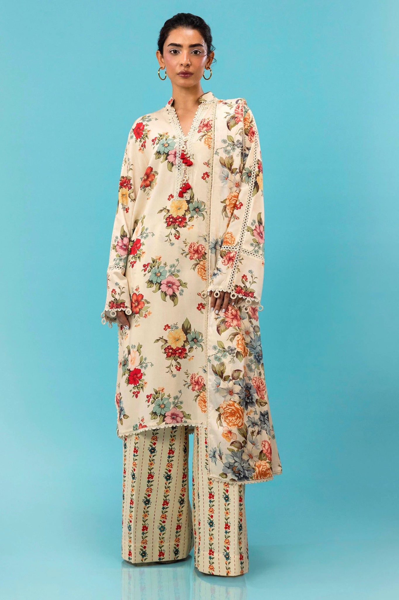 Model wearing a three - piece Sana Safinaz suit from Mahay Winter '24 featuring a cream linen shirt with vibrant floral prints, a matching printed dupatta, and striped cambric pants. Available online in the UK.