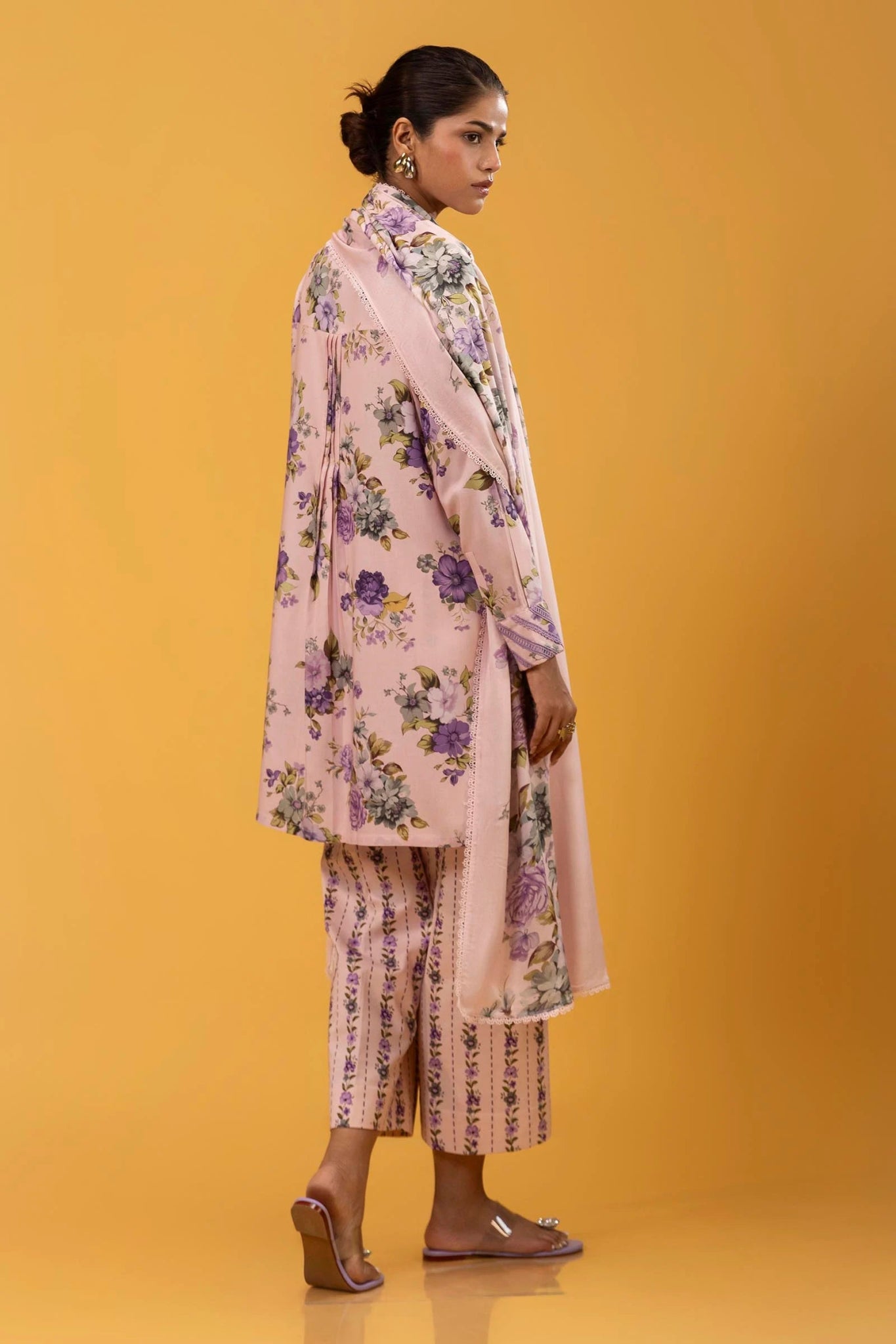 Model wearing a three - piece Sana Safinaz suit from Mahay Winter '24 in pastel pink, featuring delicate floral digital prints on a linen shirt, matching printed dupatta, and striped cambric pants. Available online in the UK.