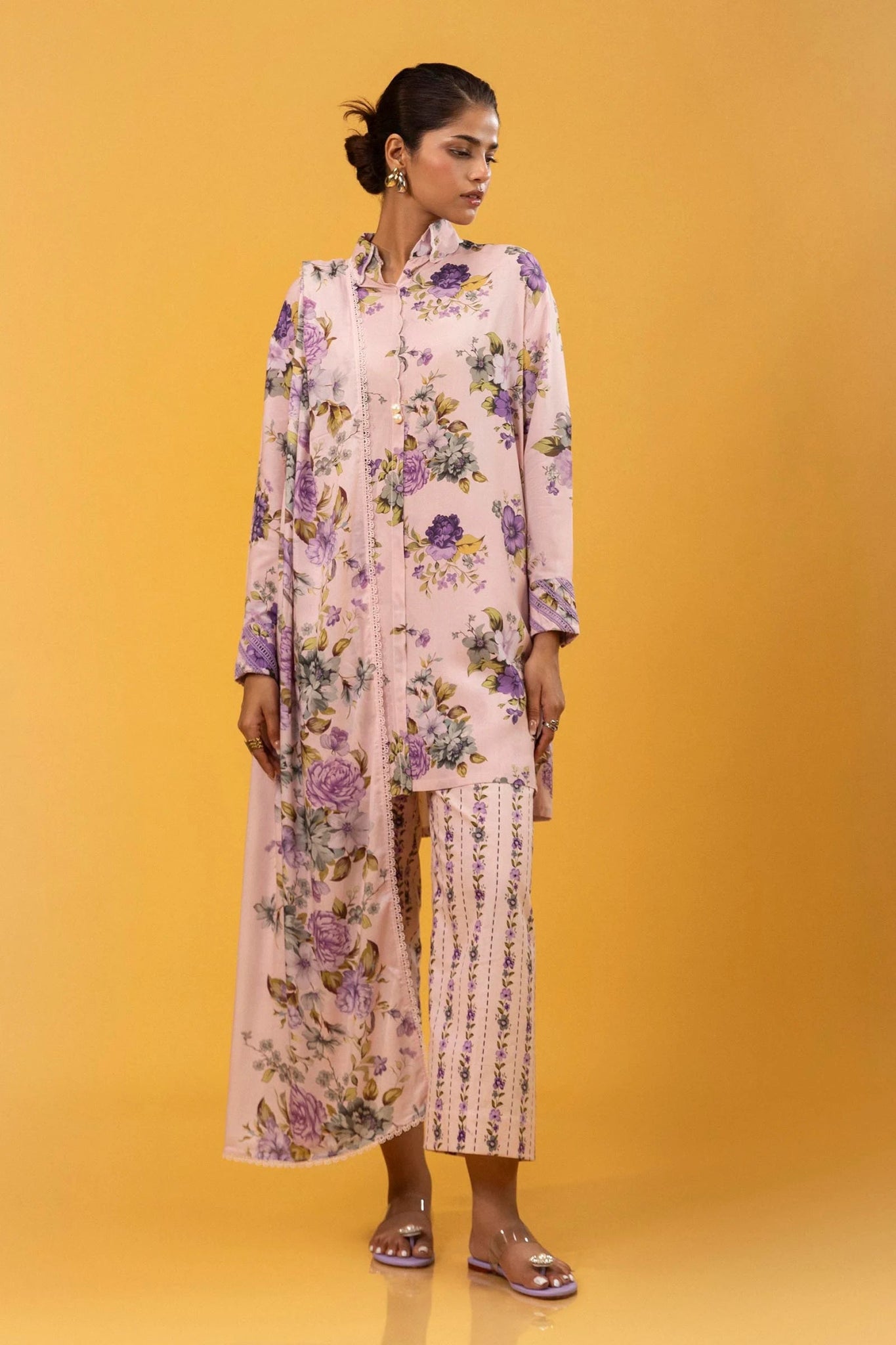 Model wearing a three - piece Sana Safinaz suit from Mahay Winter '24 in pastel pink, featuring delicate floral digital prints on a linen shirt, matching printed dupatta, and striped cambric pants. Available online in the UK.