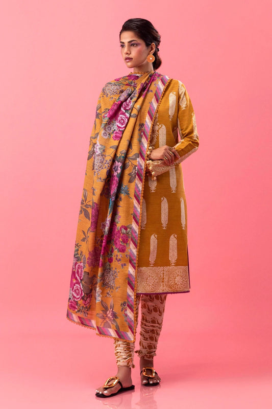 Model wearing a three - piece Sana Safinaz suit from Mahay Winter '24 in mustard yellow, featuring intricate rotary prints on slub fabric, an embroidered organza neckline, a digitally printed khaddar dupatta, and matching cambric pants. Available online in the UK.