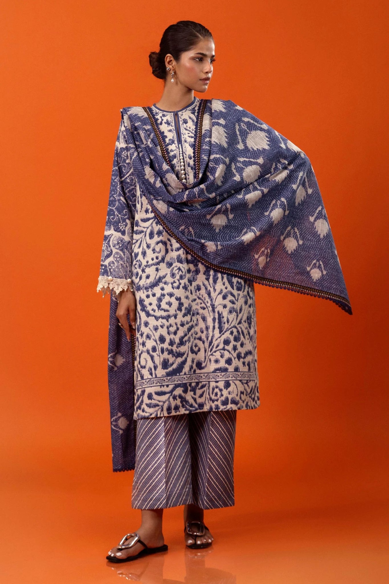 Model wearing a three - piece Sana Safinaz suit from Mahay Winter '24 in blue and cream, featuring intricate prints on slub fabric, embroidered organza detailing, a printed khaddar dupatta, and matching striped cambric pants. Available online in the UK.