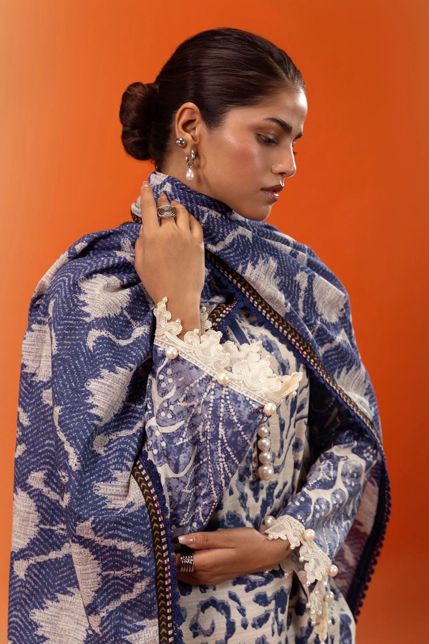 Model wearing a three - piece Sana Safinaz suit from Mahay Winter '24 in blue and cream, featuring intricate prints on slub fabric, embroidered organza detailing, a printed khaddar dupatta, and matching striped cambric pants. Available online in the UK.