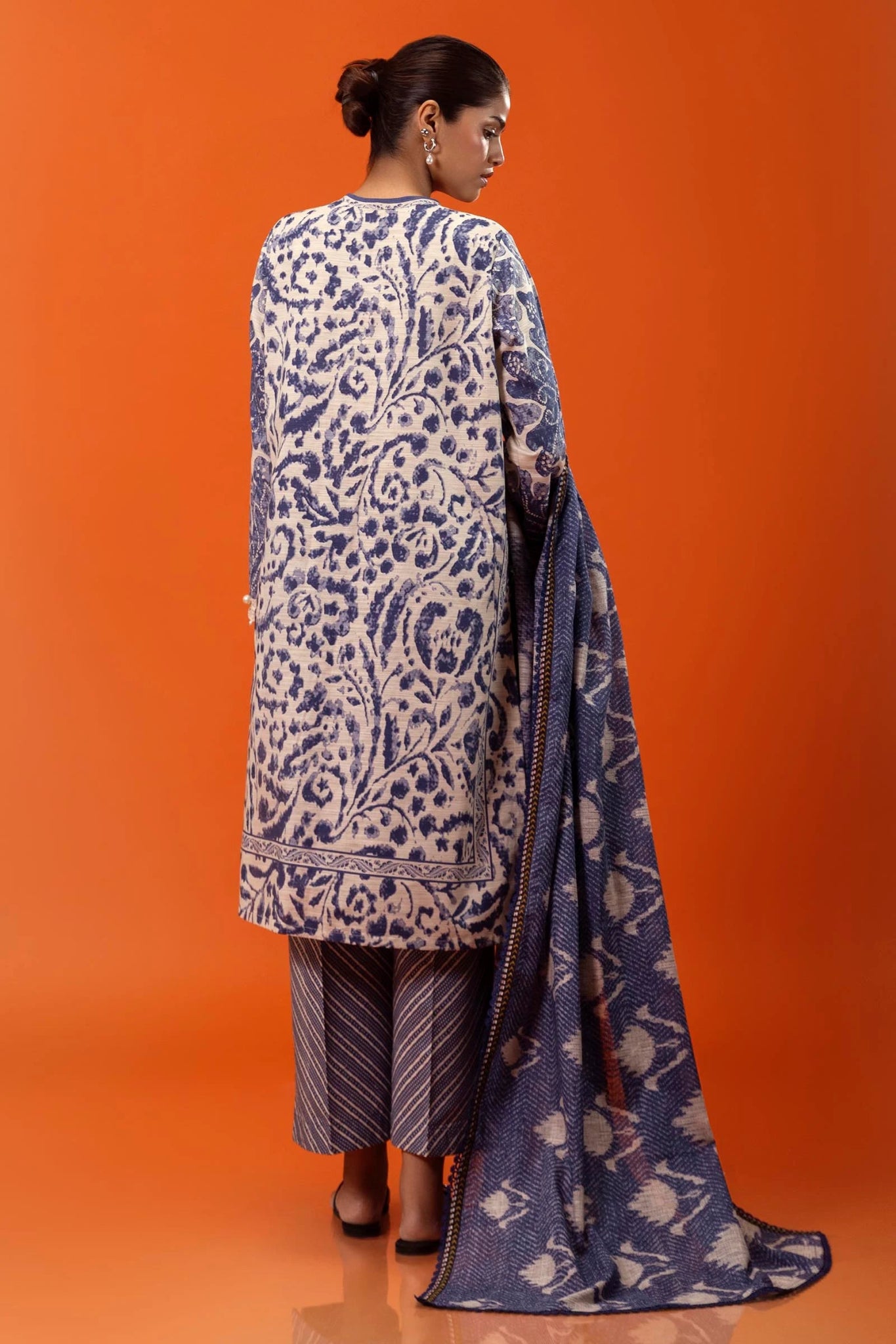 Model wearing a three - piece Sana Safinaz suit from Mahay Winter '24 in blue and cream, featuring intricate prints on slub fabric, embroidered organza detailing, a printed khaddar dupatta, and matching striped cambric pants. Available online in the UK.