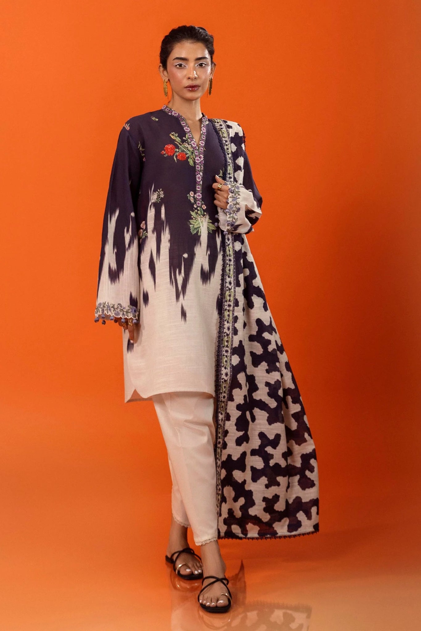 A model wearing a two - piece ensemble from Sana Safinaz, featuring a digitally printed slub shirt with floral motifs and a contrasting embroidered neckline. The look is completed with a coordinating khaddar dupatta showcasing bold, abstract patterns for a modern touch. Available online in the UK.