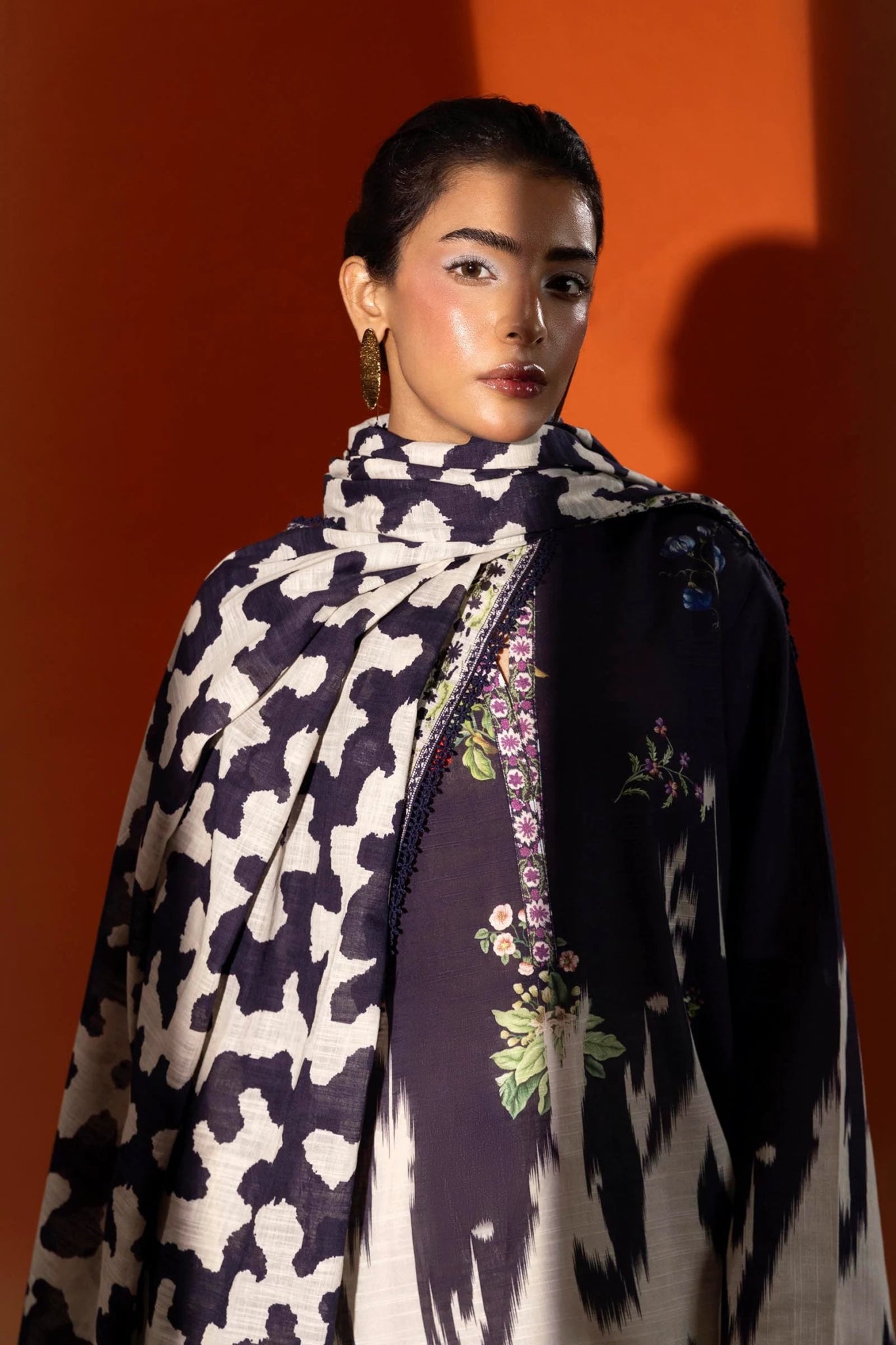 A model wearing a two - piece ensemble from Sana Safinaz, featuring a digitally printed slub shirt with floral motifs and a contrasting embroidered neckline. The look is completed with a coordinating khaddar dupatta showcasing bold, abstract patterns for a modern touch. Available online in the UK.