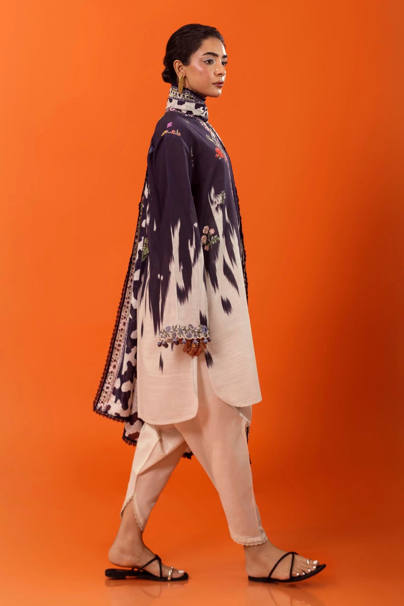 A model wearing a two - piece ensemble from Sana Safinaz, featuring a digitally printed slub shirt with floral motifs and a contrasting embroidered neckline. The look is completed with a coordinating khaddar dupatta showcasing bold, abstract patterns for a modern touch. Available online in the UK.