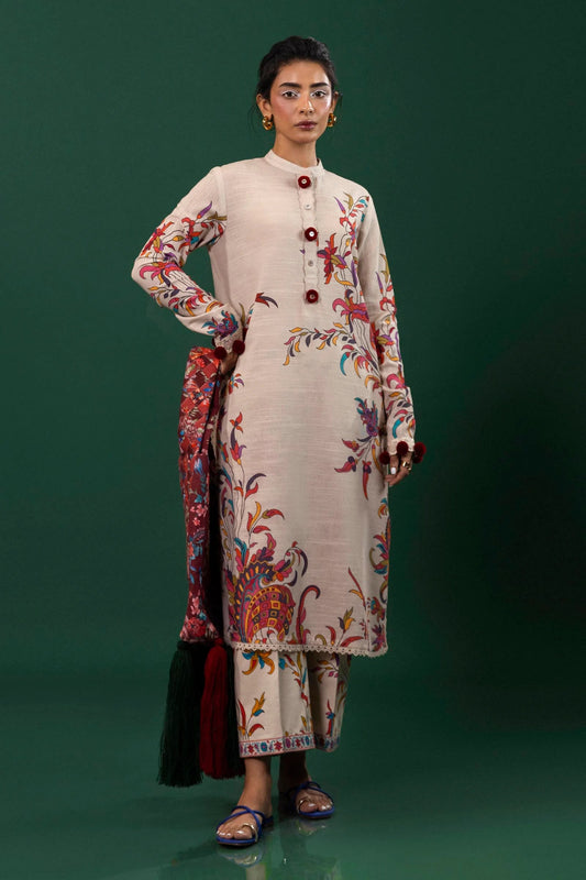 A model showcases a two - piece Sana Safinaz ensemble featuring a beige slub shirt with vibrant floral and paisley prints, accented with maroon buttons and an embroidered organza trim. The outfit includes matching printed cambric trousers, creating a cohesive look. Available online in the UK.