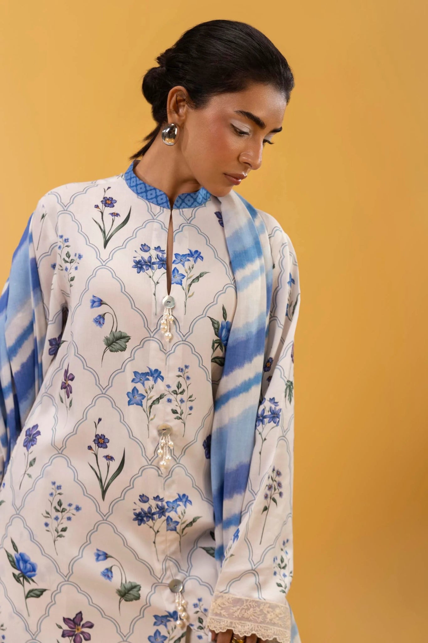 Model wearing a three - piece outfit from the Mahay Winter '24 collection by Sana Safinaz. The digitally printed linen shirt features a coordinated front, back, and sleeves with delicate embroidered patti on organza. Paired with rotary - printed cambric pants and a matching printed linen dupatta. Available online in the UK