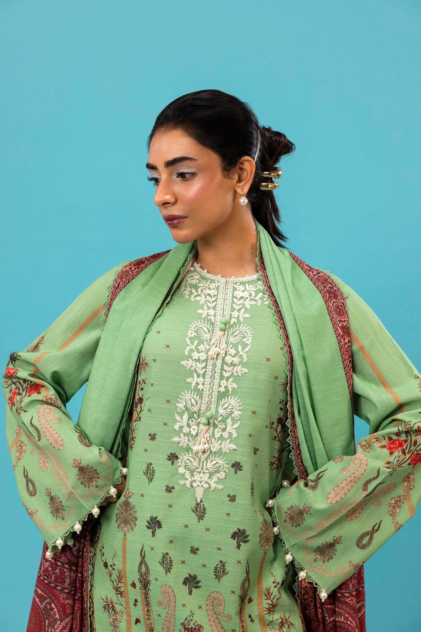 A model is wearing a three - piece outfit from the Mahay Winter '24 collection by Sana Safinaz. This ensemble includes a digitally printed slub shirt with coordinated front, back, and sleeves, and an embroidered organza neckline for added elegance. It is paired with dyed cambric pants and a digitally printed khaddar dupatta, completing the look with rich patterns. Ideal for women seeking premium Pakistani fashion, this outfit is available online in the UK for £64.00.