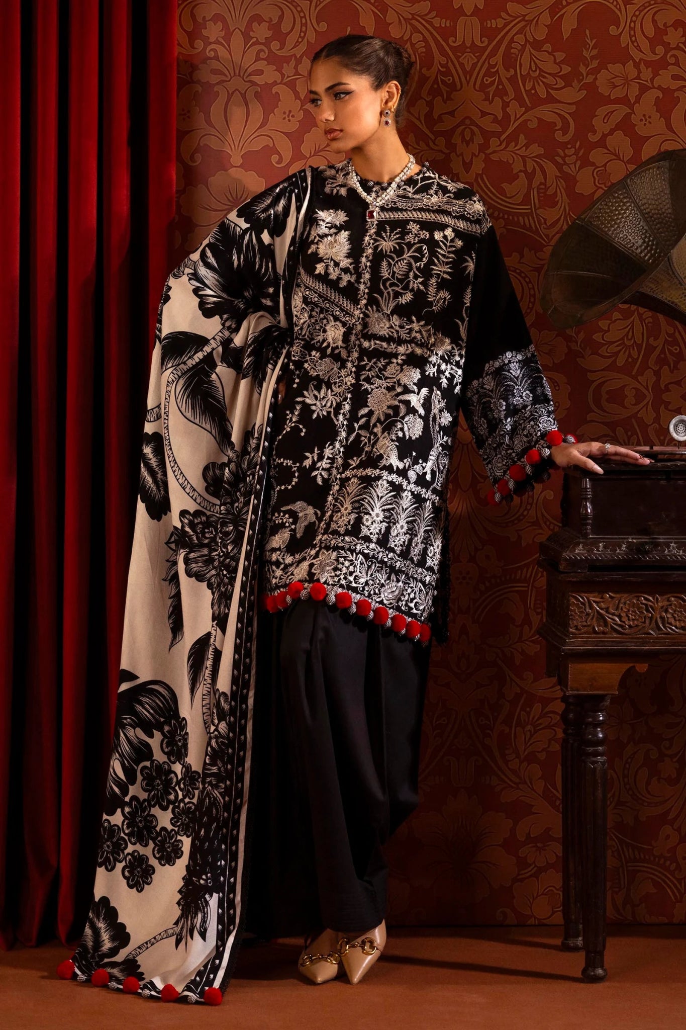 A model is wearing a three - piece outfit from the Sana Safinaz Muzlin Winter '24 Collection. This ensemble features intricate embroidered front kalis on slub fabric, a printed back, and matching sleeves. The look is completed with a beautifully printed shawl and classic dyed cotton pants. The monochrome color scheme and red accents add sophistication to this elegant Pakistani designer outfit. Available online in the UK.