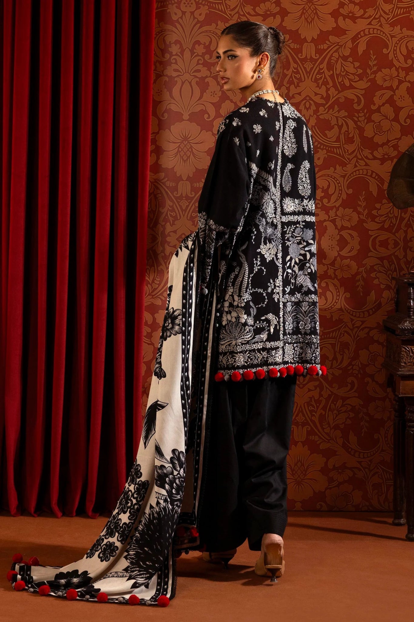 A model is wearing a three - piece outfit from the Sana Safinaz Muzlin Winter '24 Collection. This ensemble features intricate embroidered front kalis on slub fabric, a printed back, and matching sleeves. The look is completed with a beautifully printed shawl and classic dyed cotton pants. The monochrome color scheme and red accents add sophistication to this elegant Pakistani designer outfit. Available online in the UK.