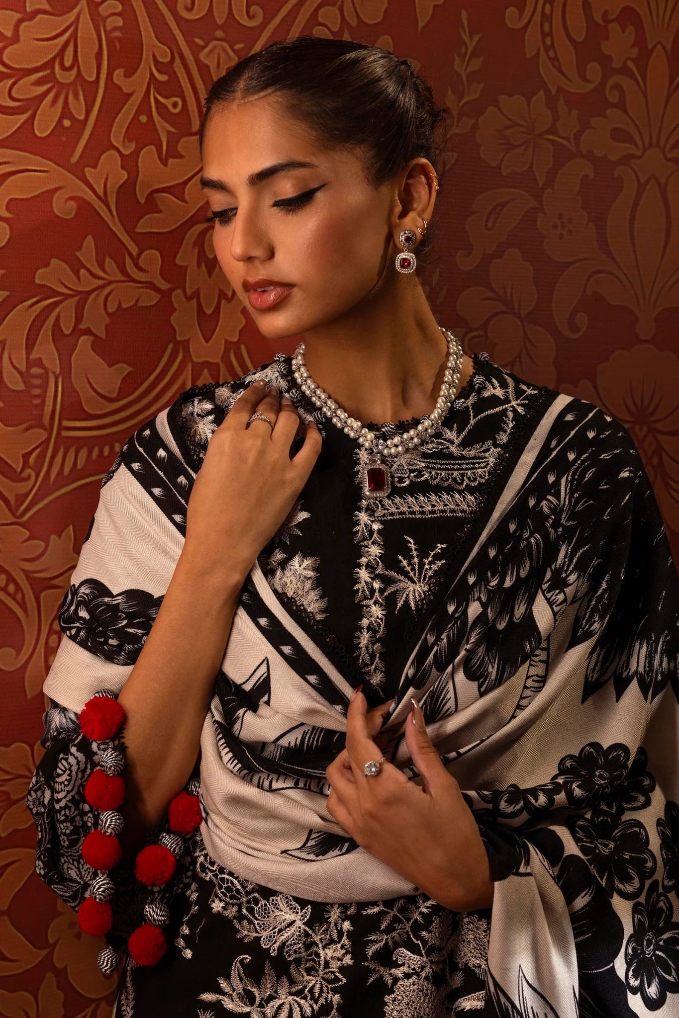 A model is wearing a three - piece outfit from the Sana Safinaz Muzlin Winter '24 Collection. This ensemble features intricate embroidered front kalis on slub fabric, a printed back, and matching sleeves. The look is completed with a beautifully printed shawl and classic dyed cotton pants. The monochrome color scheme and red accents add sophistication to this elegant Pakistani designer outfit. Available online in the UK.