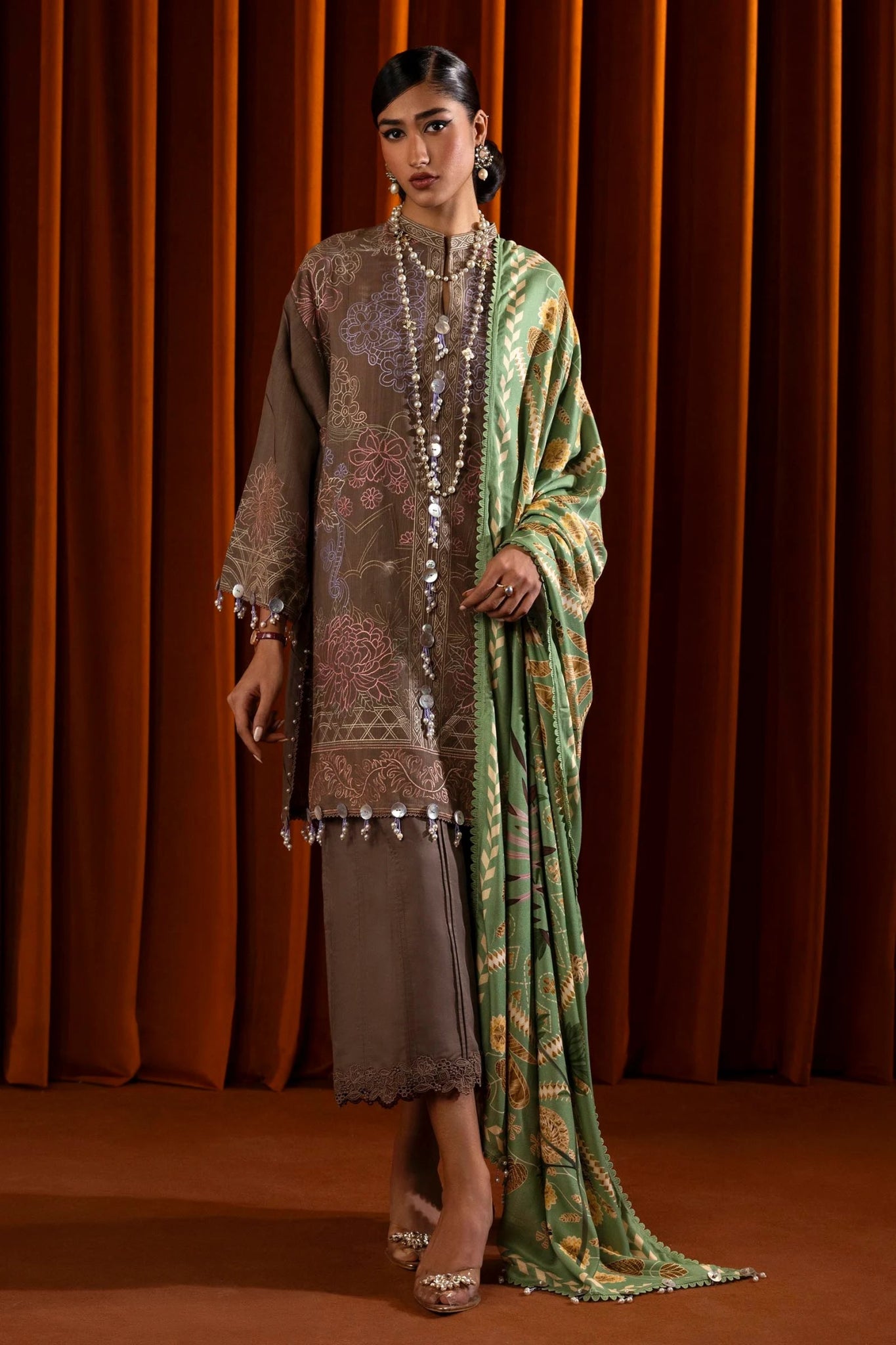 A model is wearing a three - piece outfit from the Sana Safinaz Muzlin Winter '24 Collection. The ensemble features an embroidered front kali on slub fabric, paired with a dyed slub back and sleeves, all detailed with delicate embroidery. It includes a digitally printed shawl with intricate patterns, complemented by dyed cotton pants. The luxurious and sophisticated design, with its attention to detail, offers a chic choice for women seeking elegant Pakistani designer clothing. Available online 