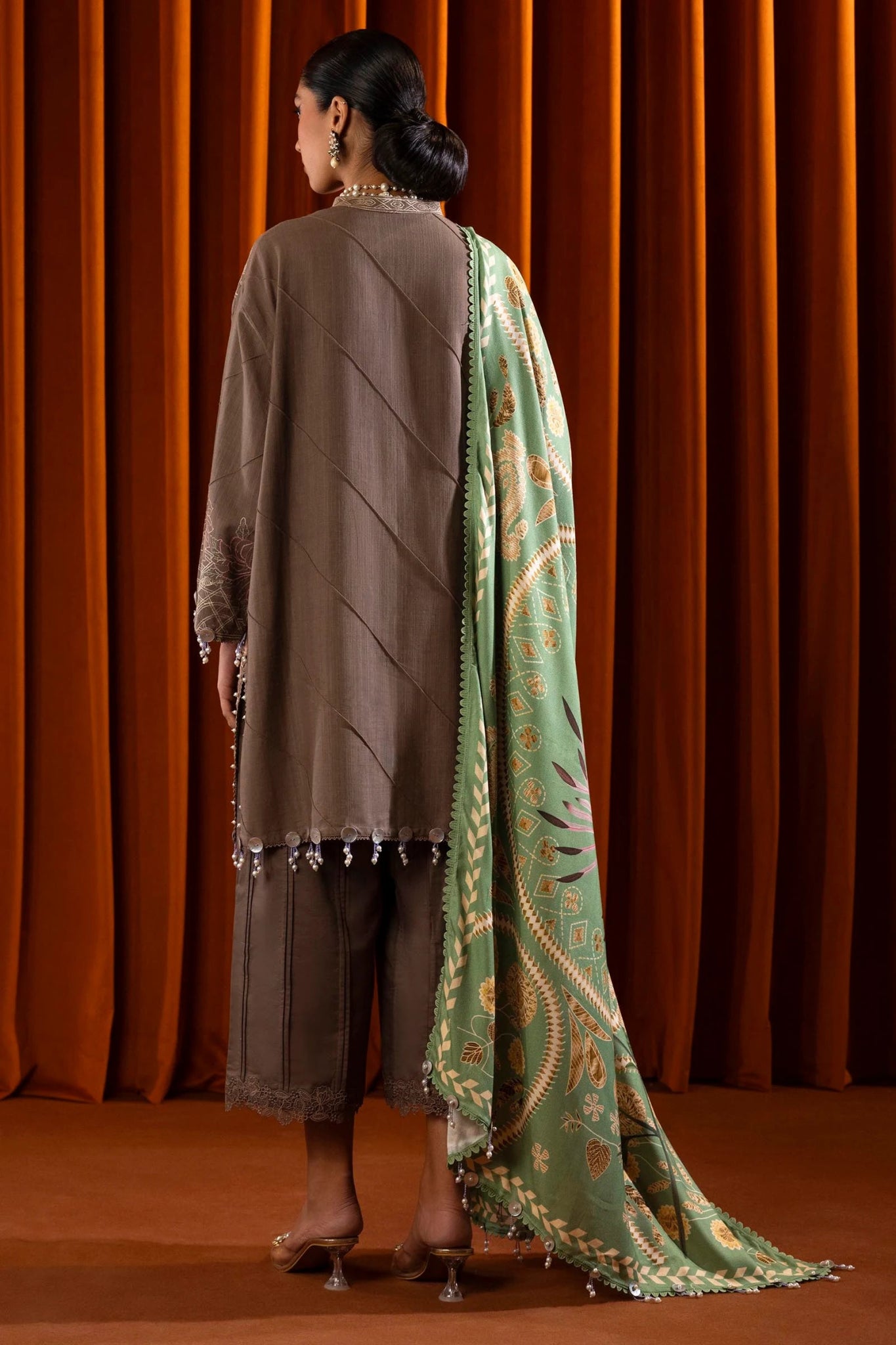 A model is wearing a three - piece outfit from the Sana Safinaz Muzlin Winter '24 Collection. The ensemble features an embroidered front kali on slub fabric, paired with a dyed slub back and sleeves, all detailed with delicate embroidery. It includes a digitally printed shawl with intricate patterns, complemented by dyed cotton pants. The luxurious and sophisticated design, with its attention to detail, offers a chic choice for women seeking elegant Pakistani designer clothing. Available online 