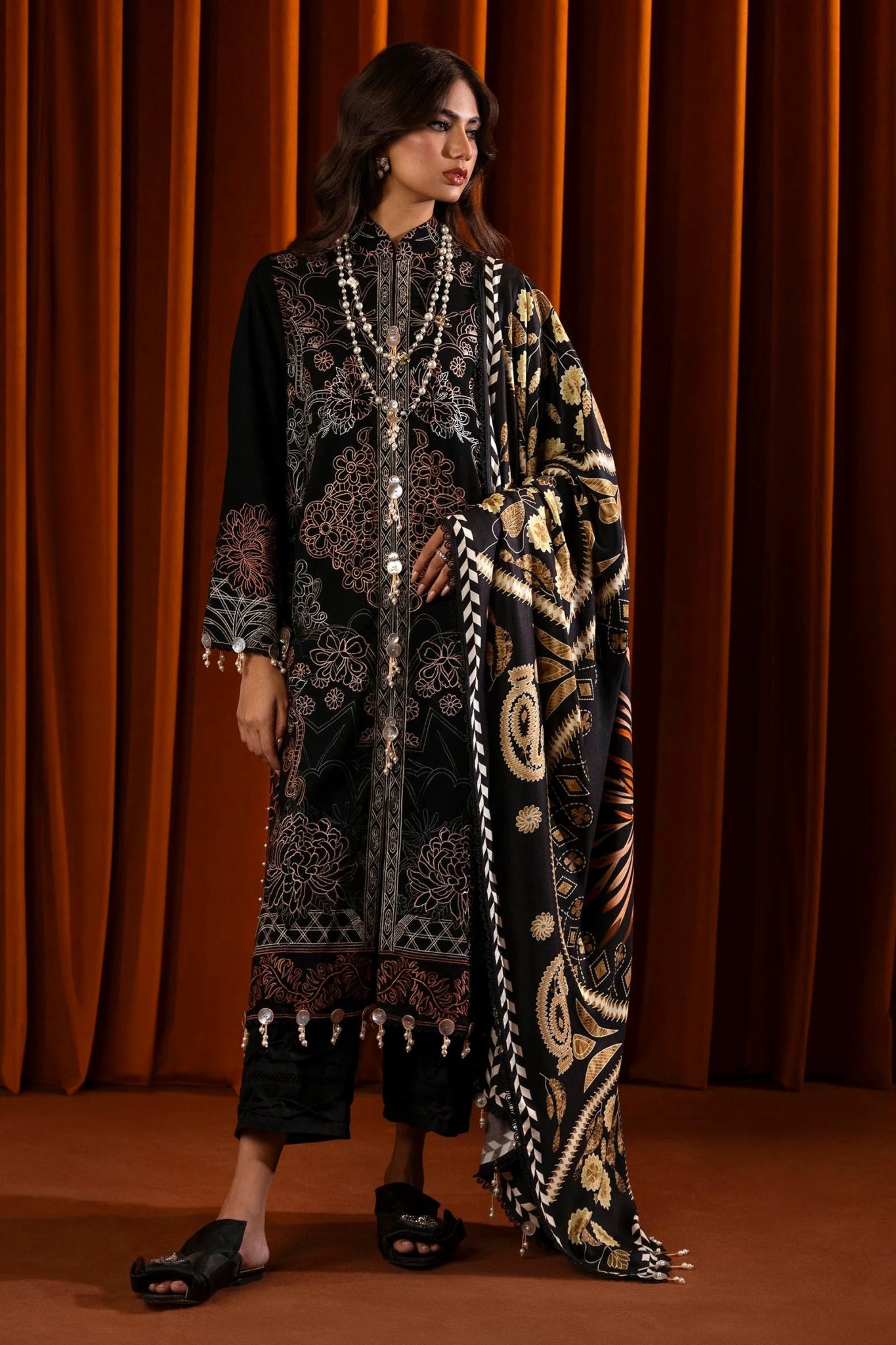 A model wears a three - piece outfit from the Sana Safinaz Muzlin Winter '24 Collection. The ensemble features an embroidered slub shirt with front kali panels, dyed slub back, and embroidered sleeves. Paired with a digitally printed shawl showcasing intricate patterns and dyed cotton pants, this luxurious Pakistani outfit blends traditional embroidery with contemporary design.