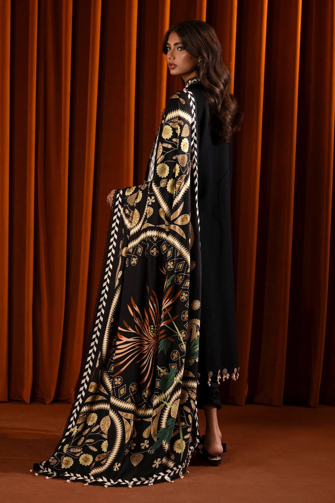 A model wears a three - piece outfit from the Sana Safinaz Muzlin Winter '24 Collection. The ensemble features an embroidered slub shirt with front kali panels, dyed slub back, and embroidered sleeves. Paired with a digitally printed shawl showcasing intricate patterns and dyed cotton pants, this luxurious Pakistani outfit blends traditional embroidery with contemporary design.