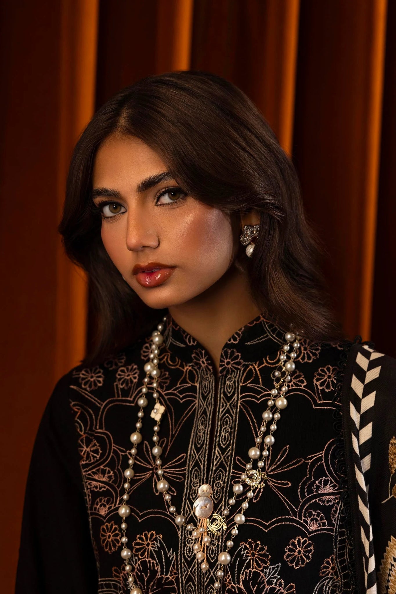 A model wears a three - piece outfit from the Sana Safinaz Muzlin Winter '24 Collection. The ensemble features an embroidered slub shirt with front kali panels, dyed slub back, and embroidered sleeves. Paired with a digitally printed shawl showcasing intricate patterns and dyed cotton pants, this luxurious Pakistani outfit blends traditional embroidery with contemporary design.