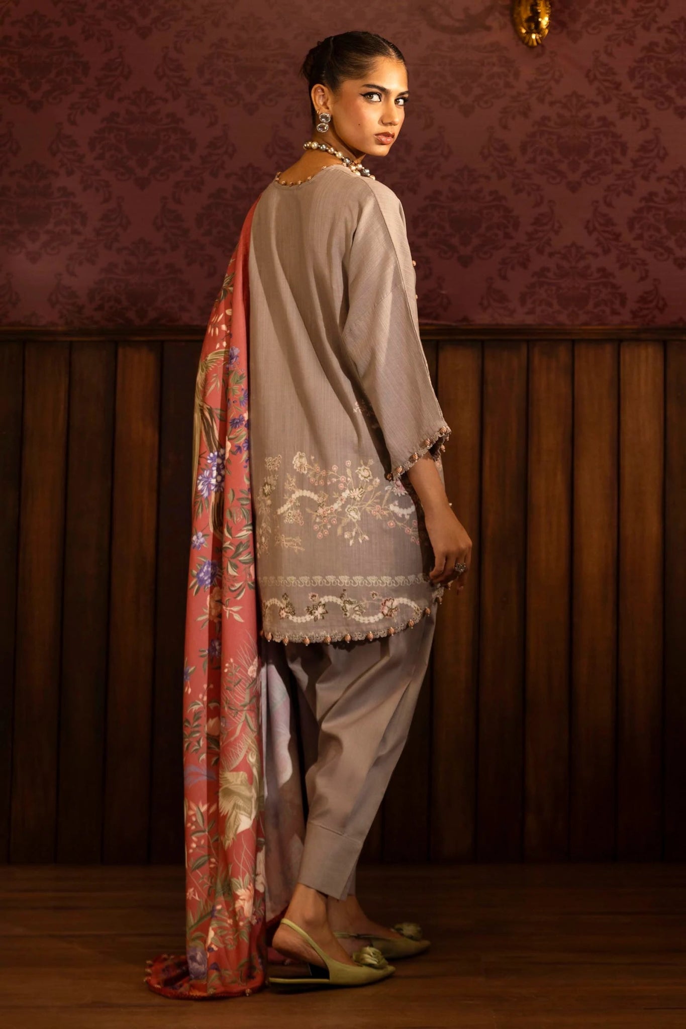 A model wears a three - piece outfit from the Sana Safinaz Muzlin Winter '24 Collection. It features an embroidered front kali on slub, printed back, and embroidered sleeves, paired with a vibrant, digitally printed shawl and matching dyed cotton pants. This elegant look blends traditional embroidery with a modern flair, ideal for women seeking premium Pakistani fashion online in the UK.