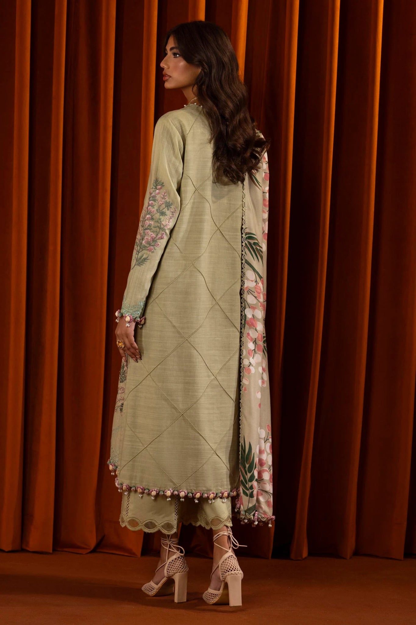 A model wears a three - piece outfit from the Sana Safinaz Muzlin Winter '24 Collection. This ensemble includes an embroidered front kali on slub fabric, a dyed back, and embroidered sleeves with organza kuful details. The look is complemented by a digital printed shawl and dyed cotton pants, offering an elegant and cohesive style. Perfect for women seeking luxurious Pakistani designer clothing in the UK.