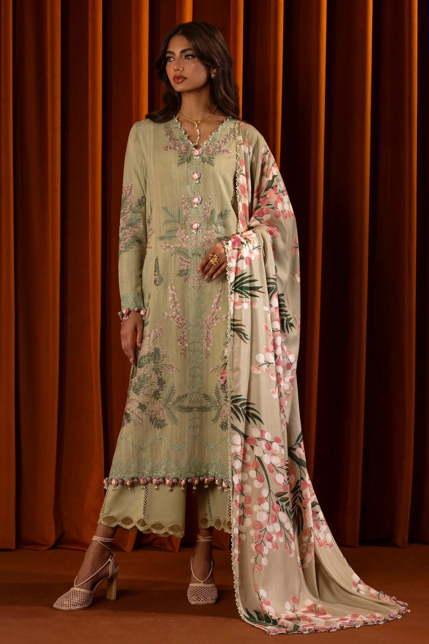 A model wears a three - piece outfit from the Sana Safinaz Muzlin Winter '24 Collection. This ensemble includes an embroidered front kali on slub fabric, a dyed back, and embroidered sleeves with organza kuful details. The look is complemented by a digital printed shawl and dyed cotton pants, offering an elegant and cohesive style. Perfect for women seeking luxurious Pakistani designer clothing in the UK.