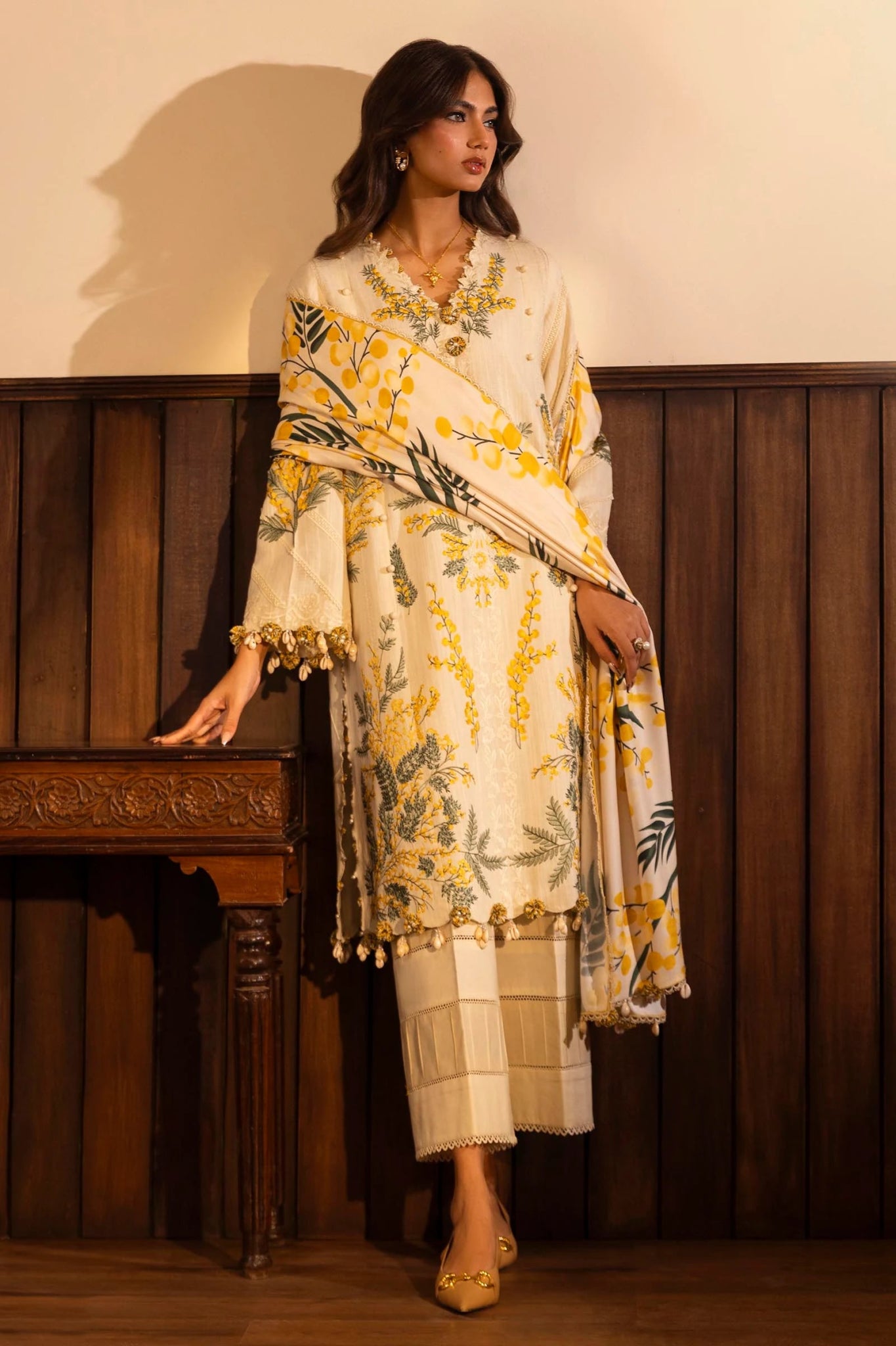 A model is wearing a three - piece outfit from the Sana Safinaz Muzlin Winter '24 Collection. The ensemble features an embroidered front kali on slub fabric, paired with a dyed slub back and sleeves, accentuated by organza embroidered kuful. The look is completed with a vibrant, digitally printed shawl and dyed cotton pants, combining traditional elegance with a modern aesthetic. Perfect for women seeking luxurious Pakistani designer clothing in the UK.