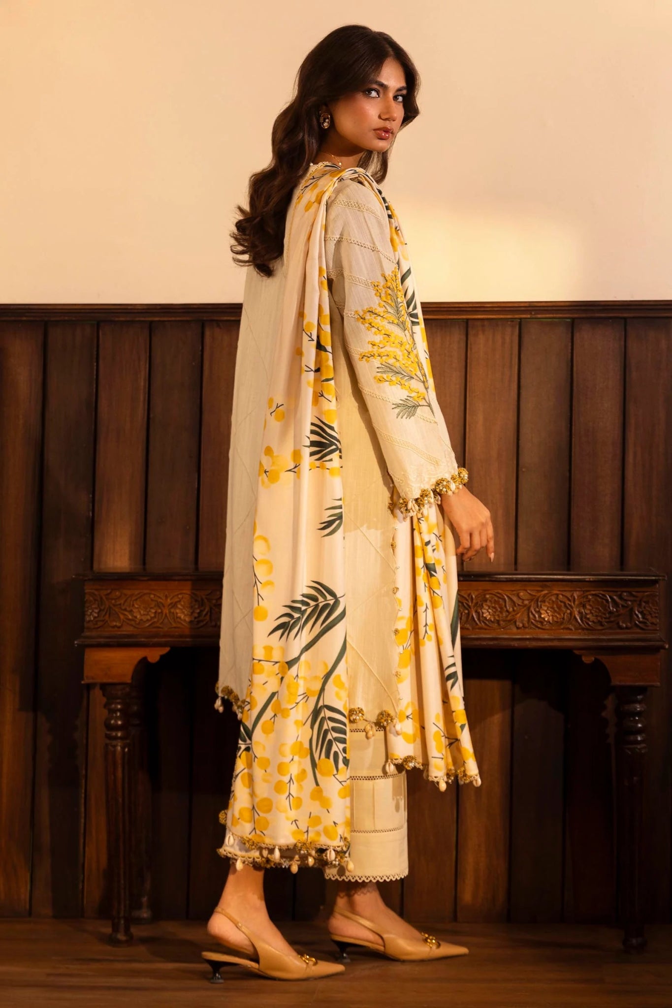 A model is wearing a three - piece outfit from the Sana Safinaz Muzlin Winter '24 Collection. The ensemble features an embroidered front kali on slub fabric, paired with a dyed slub back and sleeves, accentuated by organza embroidered kuful. The look is completed with a vibrant, digitally printed shawl and dyed cotton pants, combining traditional elegance with a modern aesthetic. Perfect for women seeking luxurious Pakistani designer clothing in the UK.