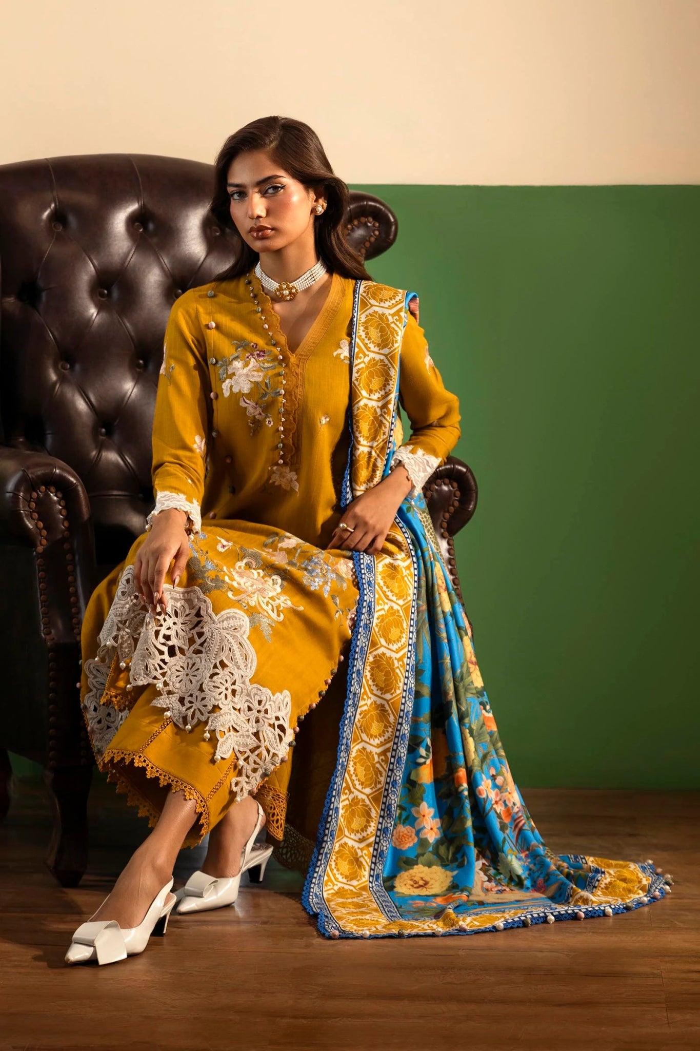 A model wears a mustard - colored three - piece outfit from the Sana Safinaz Muzlin Winter '24 Collection. The set features embroidered front panels on slub fabric, a dyed slub back, and embroidered sleeves. It includes a delicately embroidered organza border and patti, adding refined detailing. Paired with a vibrant digitally printed shawl and dyed cotton pants, the ensemble offers a sophisticated and stylish look perfect for Pakistani designer wear.