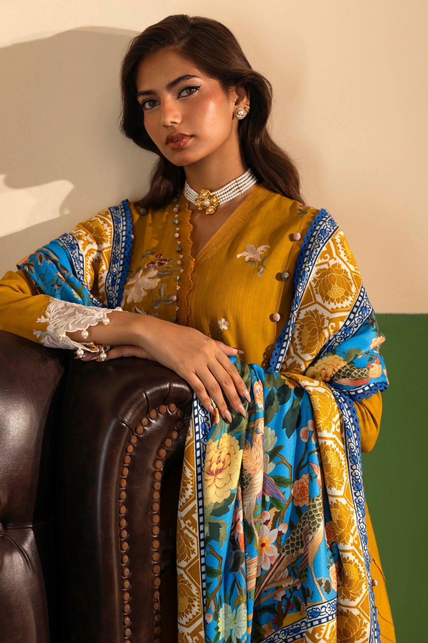 A model wears a mustard - colored three - piece outfit from the Sana Safinaz Muzlin Winter '24 Collection. The set features embroidered front panels on slub fabric, a dyed slub back, and embroidered sleeves. It includes a delicately embroidered organza border and patti, adding refined detailing. Paired with a vibrant digitally printed shawl and dyed cotton pants, the ensemble offers a sophisticated and stylish look perfect for Pakistani designer wear.