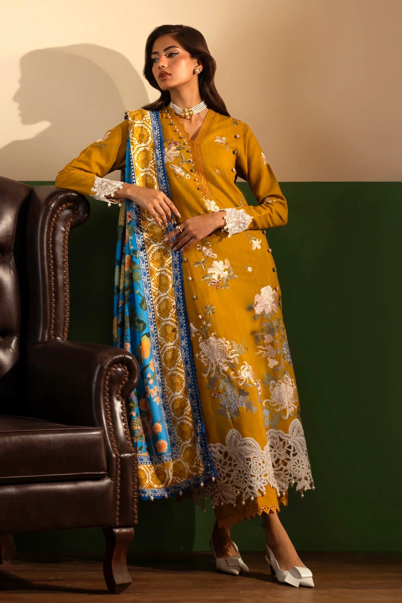 A model wears a mustard - colored three - piece outfit from the Sana Safinaz Muzlin Winter '24 Collection. The set features embroidered front panels on slub fabric, a dyed slub back, and embroidered sleeves. It includes a delicately embroidered organza border and patti, adding refined detailing. Paired with a vibrant digitally printed shawl and dyed cotton pants, the ensemble offers a sophisticated and stylish look perfect for Pakistani designer wear.