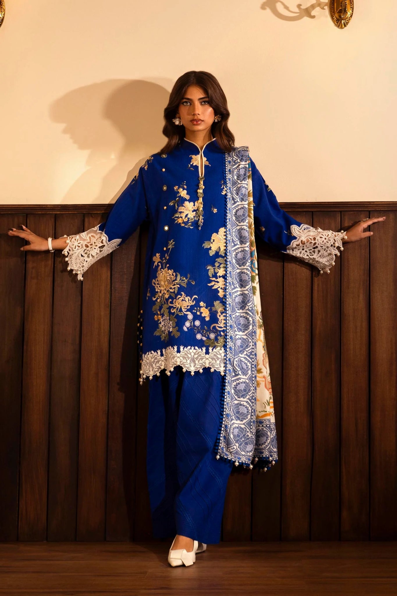 A model wears a three - piece outfit from the Sana Safinaz Muzlin Winter '24 Collection. The ensemble features an embroidered slub front kali with intricate detailing on the sleeves and borders. It includes a vibrant digital - printed shawl and dyed cotton pants for a complete look. Ideal for women seeking luxurious and elegant Pakistani fashion.