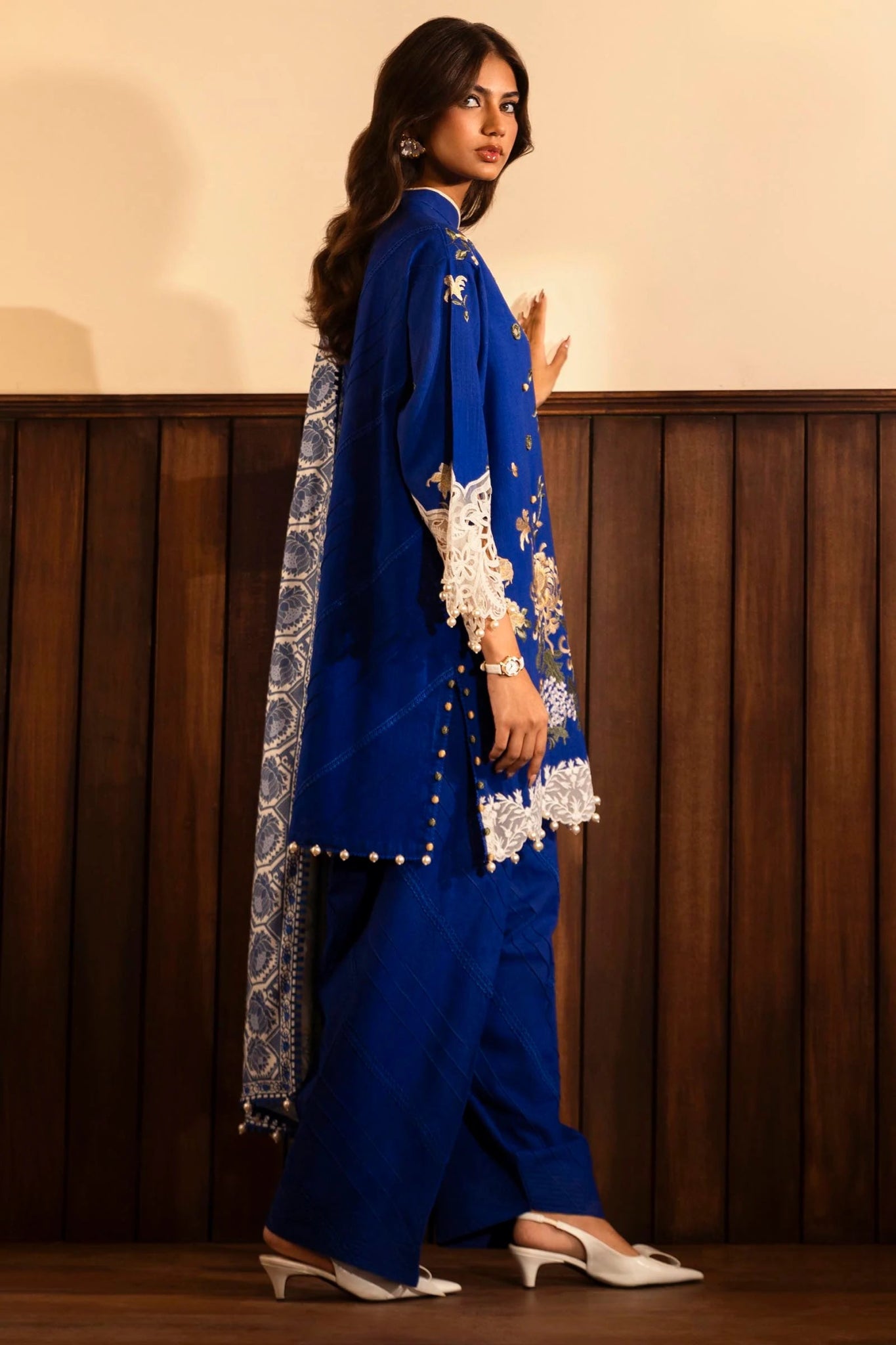 A model wears a three - piece outfit from the Sana Safinaz Muzlin Winter '24 Collection. The ensemble features an embroidered slub front kali with intricate detailing on the sleeves and borders. It includes a vibrant digital - printed shawl and dyed cotton pants for a complete look. Ideal for women seeking luxurious and elegant Pakistani fashion.